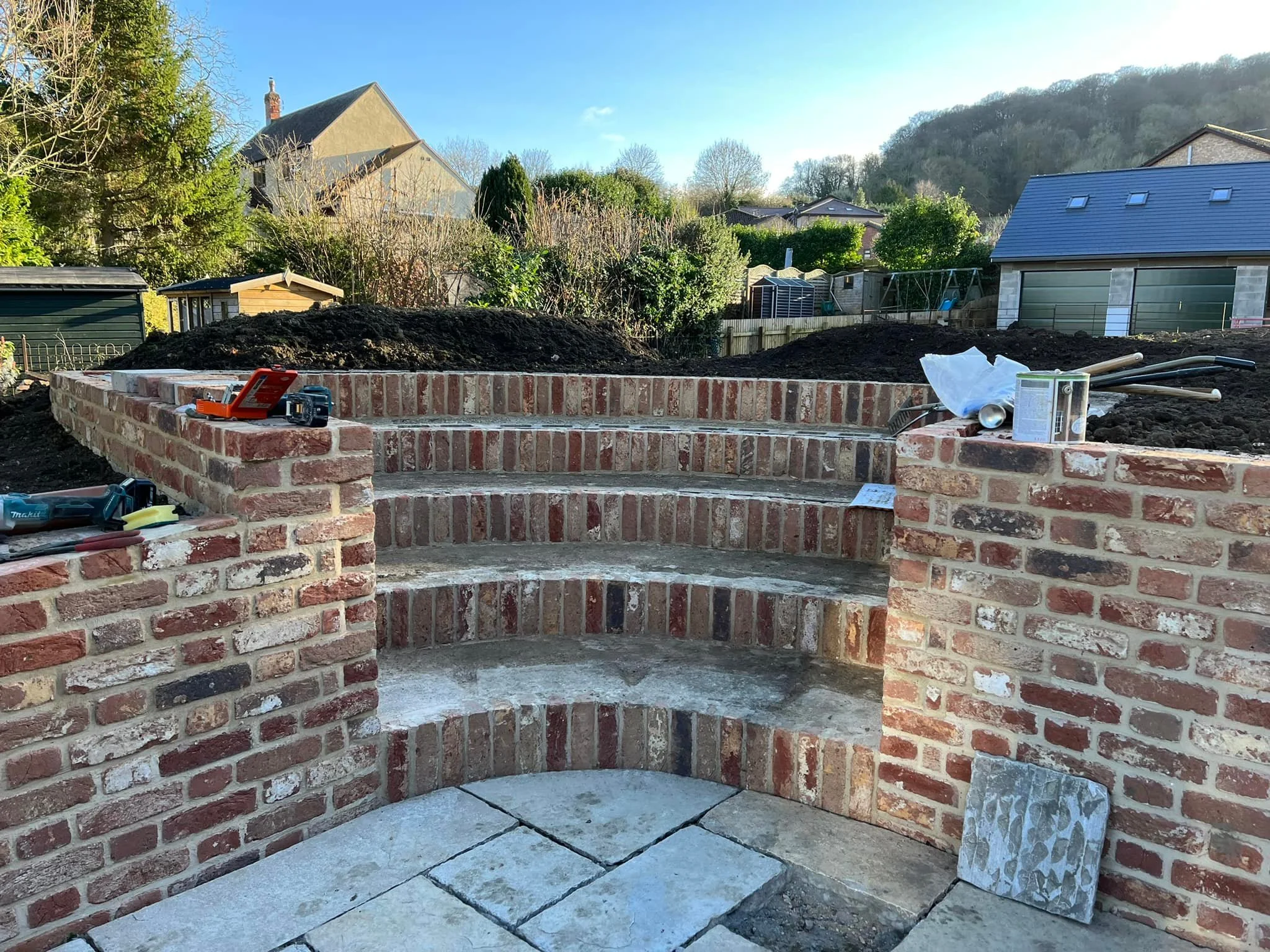 Brick staircase under construction outdoors with tools and materials on top, surrounded by soil and garden in the background.