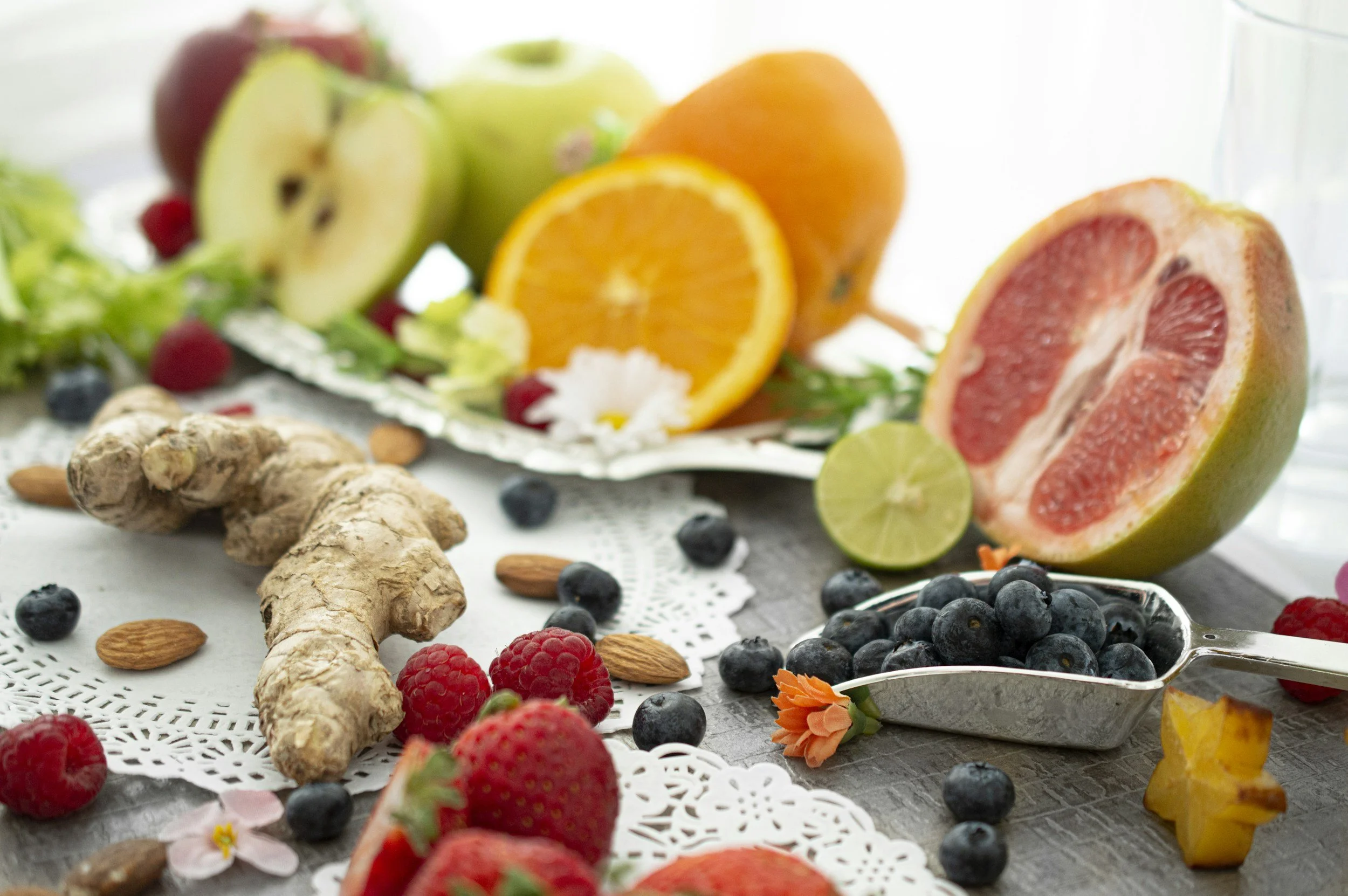 Assorted fresh fruits including grapefruit, oranges, apples, blueberries, raspberries, lemon, and lime, along with ginger and almonds, arranged on a table with decorative doilies and flowers.