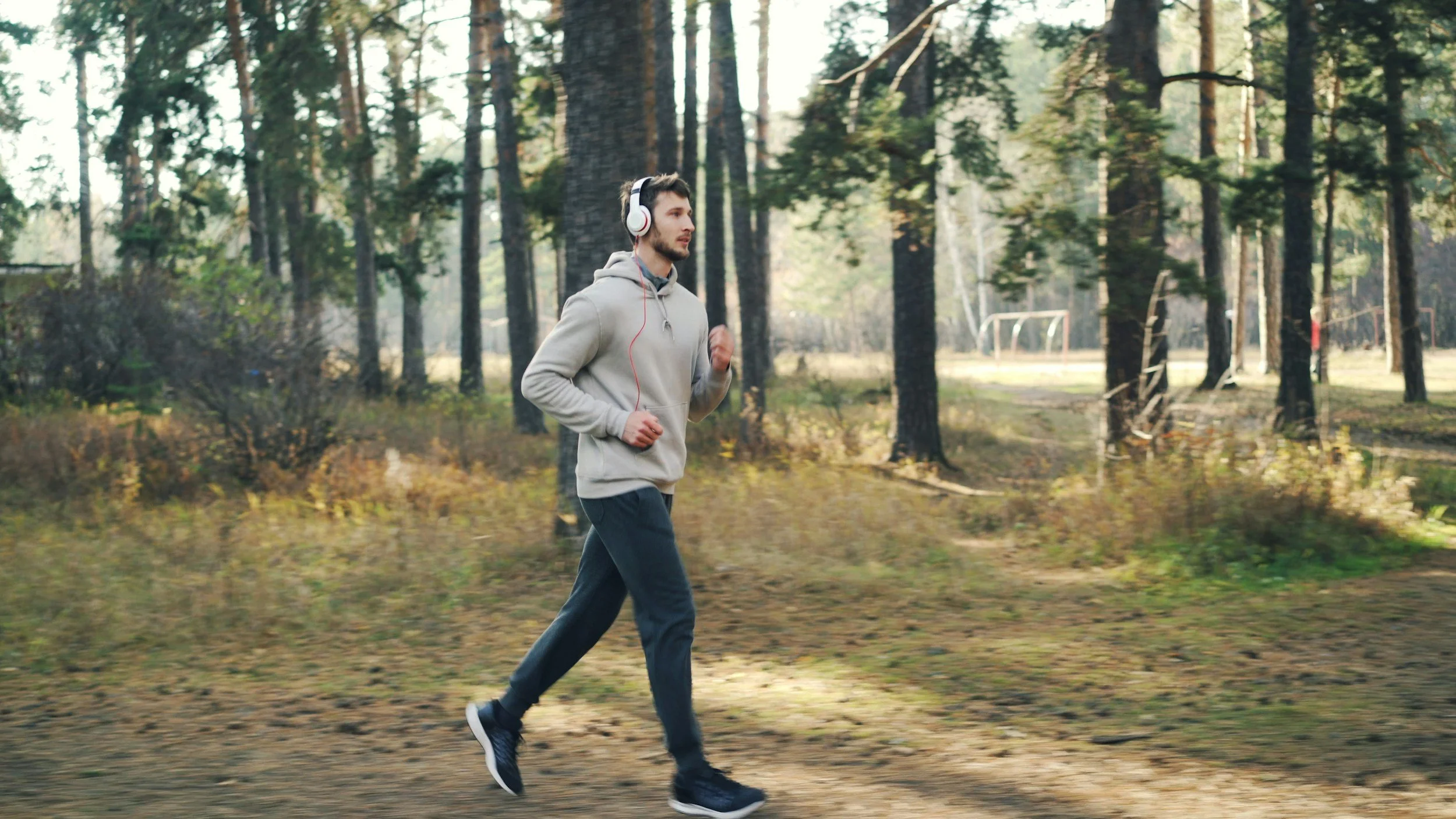 A young man jogging through a wooded park with tall trees, wearing a light gray hoodie, black pants, and headphones.