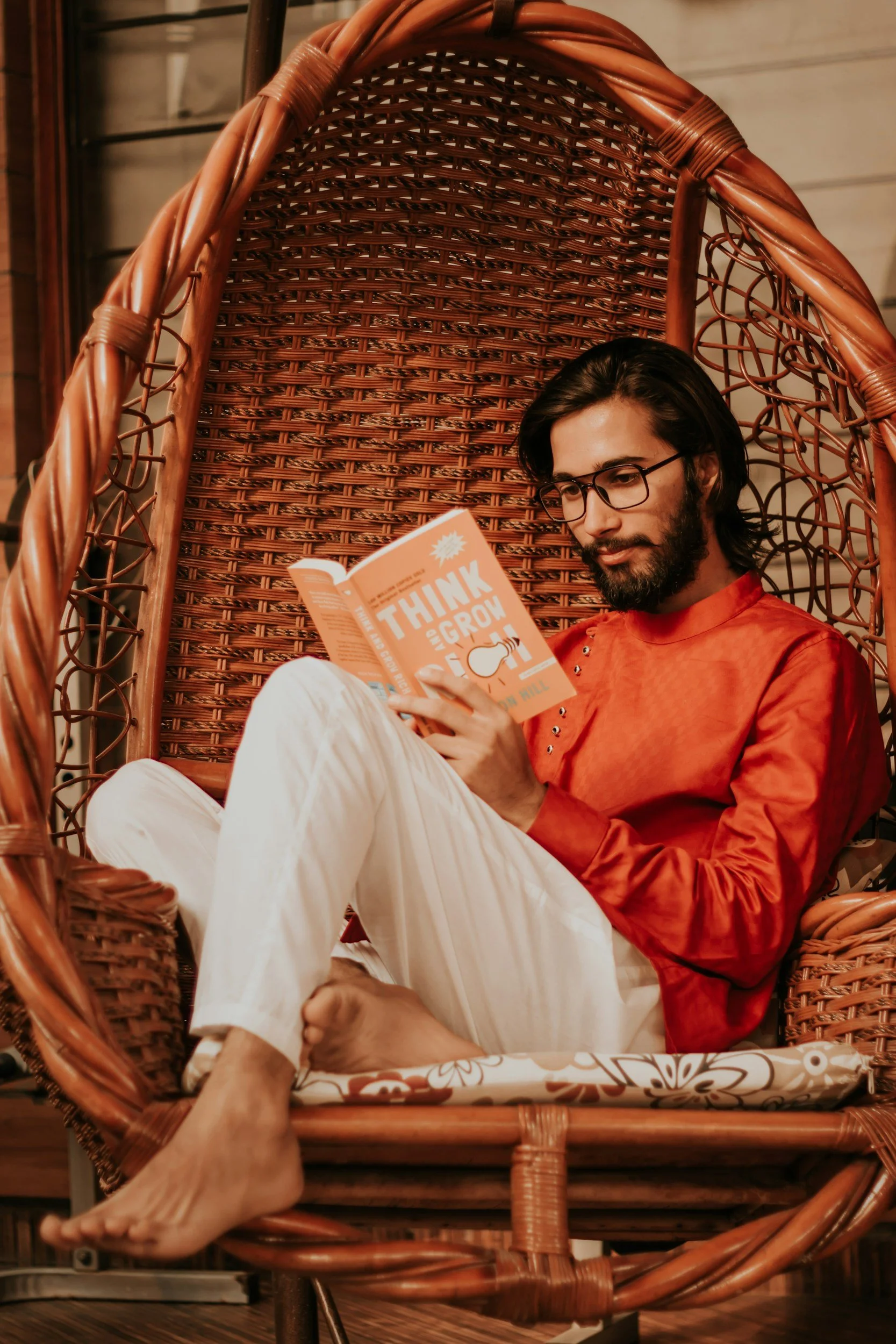 A man with dark hair, glasses, and a beard is sitting in a large wicker chair, reading a book titled "Think and Grow." He is wearing a red shirt and white pants, with his legs crossed and barefoot in a cozy indoor setting.