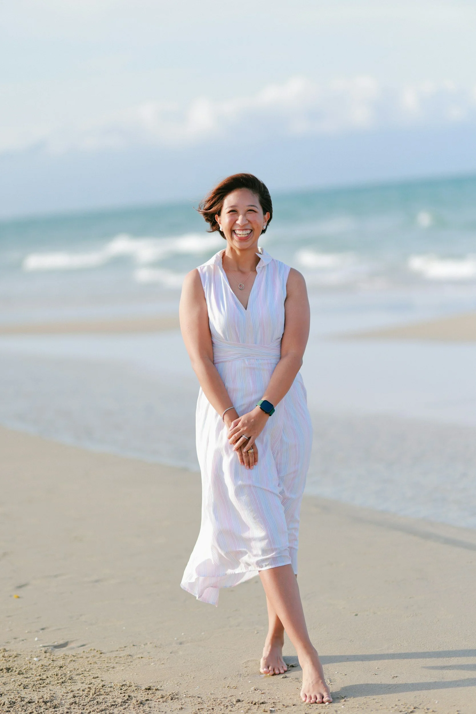 A woman in a white dress walking barefoot on a beach with the ocean and cloudy sky in the background, smiling joyfully.