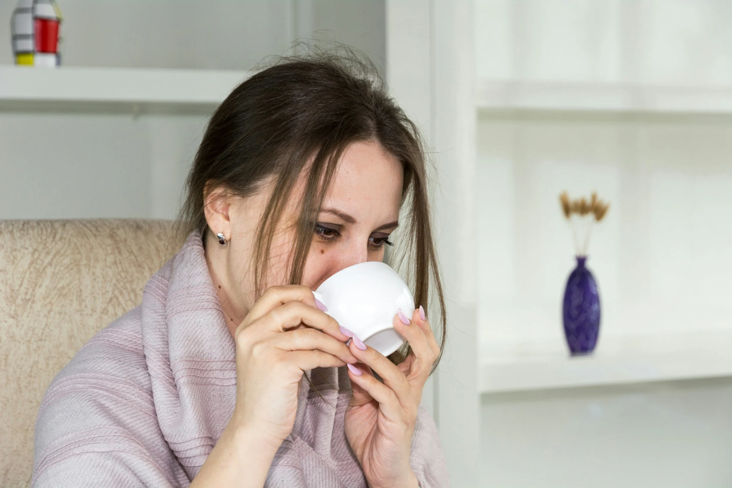 A woman sitting in a beige chair drinking from a white mug in a bright room with white shelves and a purple vase with dried flowers in the background.