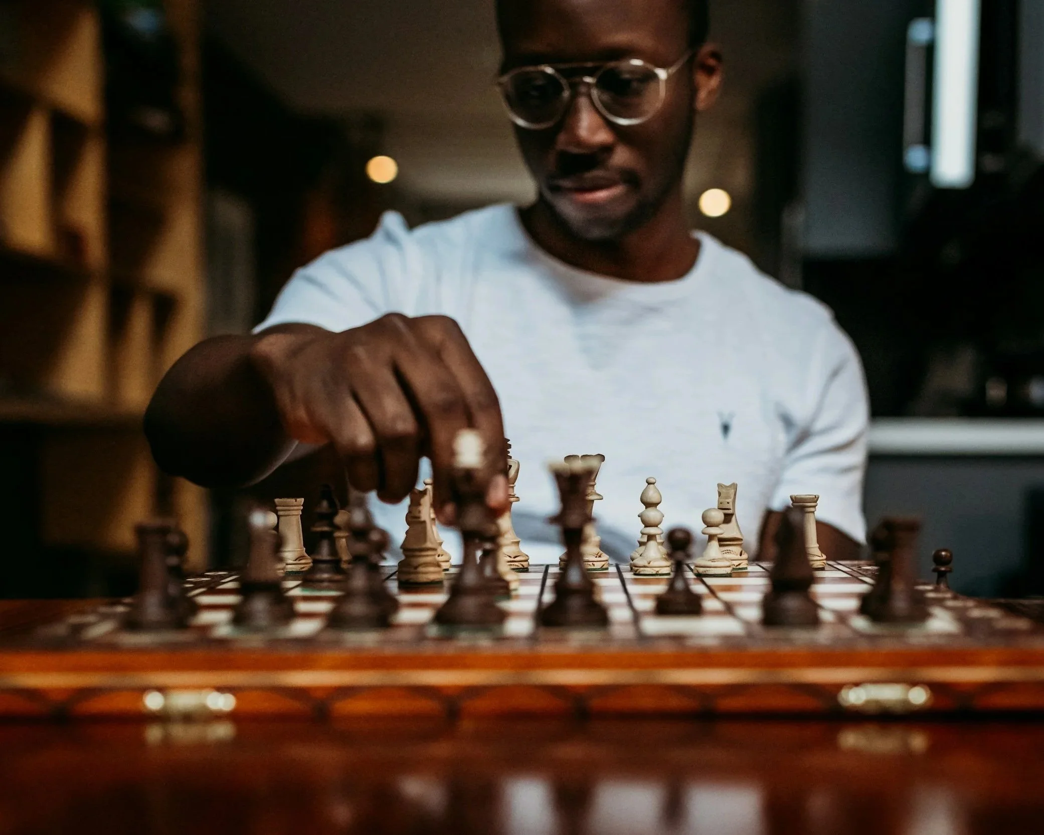 A man wearing glasses and a white t-shirt playing chess indoors.