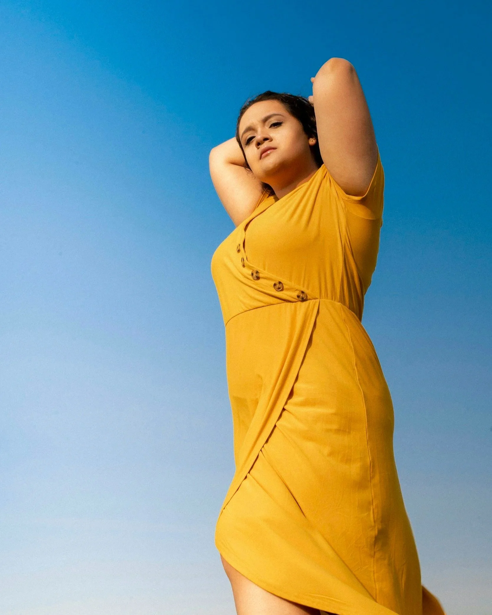 A woman in a yellow dress standing outdoors against a clear blue sky, with her arms raised behind her head.
