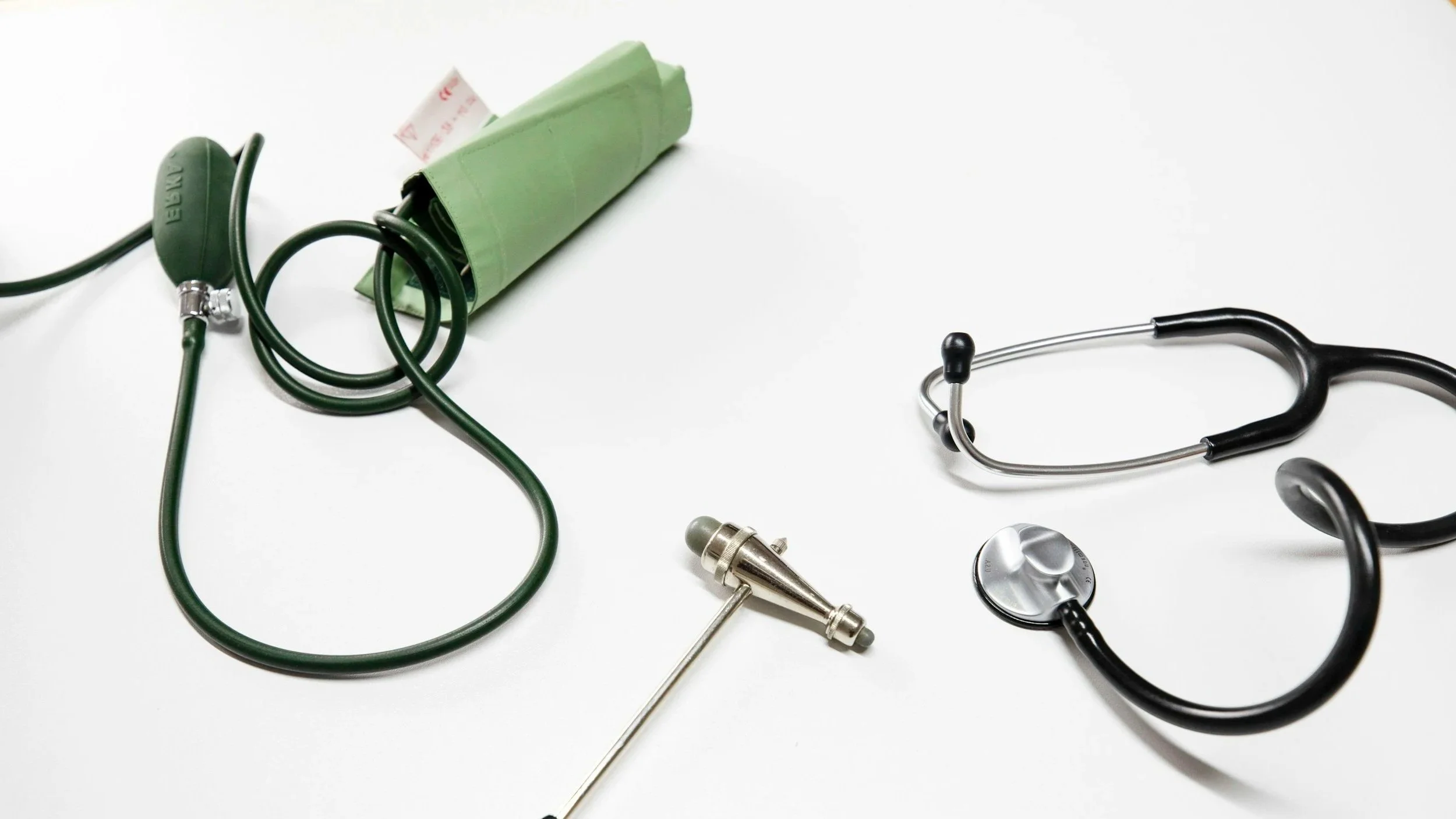 Medical equipment including a green blood pressure cuff, a green blood pressure bulb, a metal syringe, and black stethoscopes on a white background.
