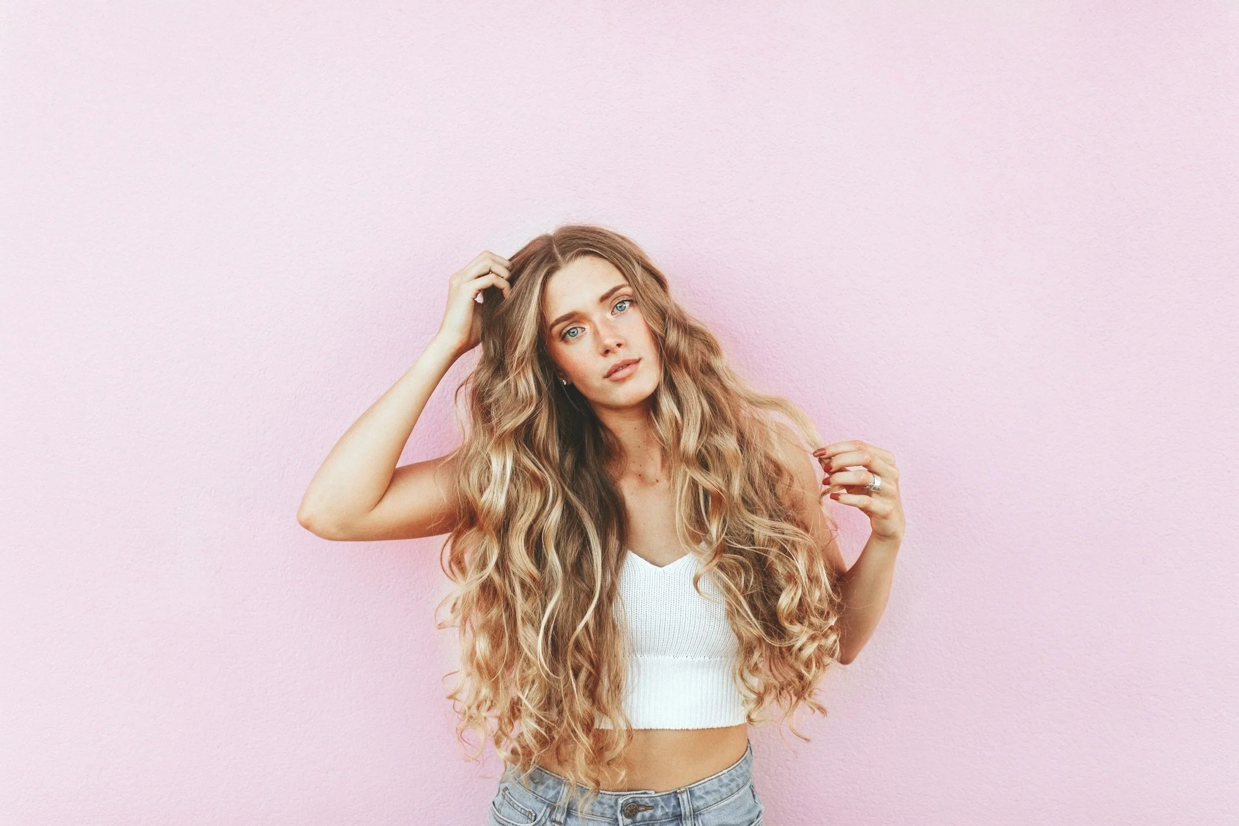 Young woman with long, wavy blonde hair wearing a white sleeveless top and denim jeans standing against a pink wall.