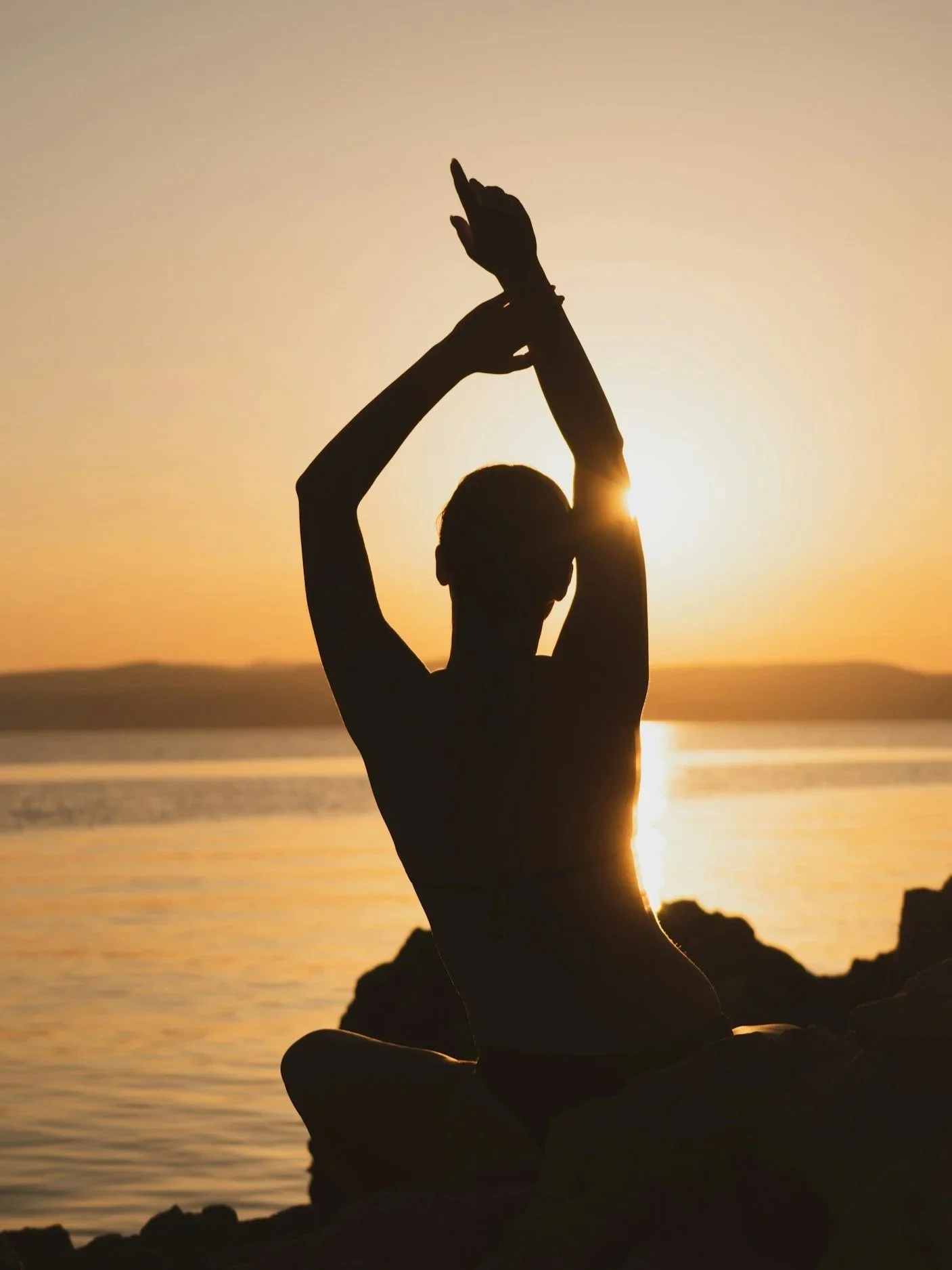 Silhouette of a woman practicing yoga or meditation at sunset on a rocky beach.