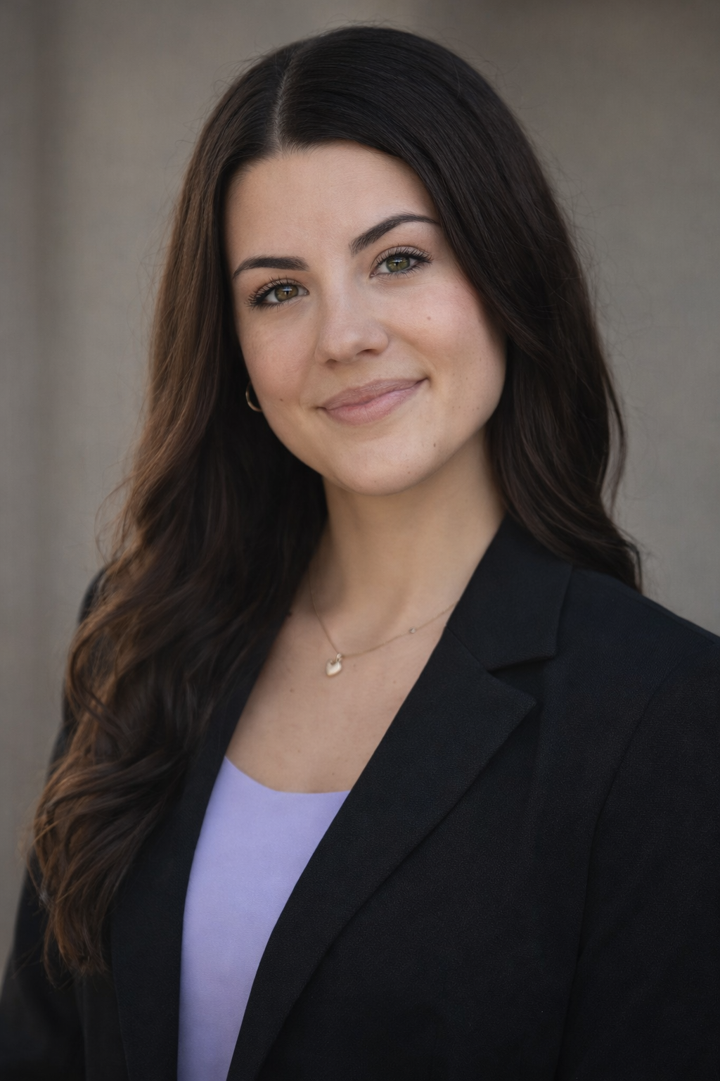 A young woman with long, wavy dark hair and light skin, smiling, wearing a black blazer over a lavender top, with a small heart-shaped necklace, standing outdoors against a gray concrete background.