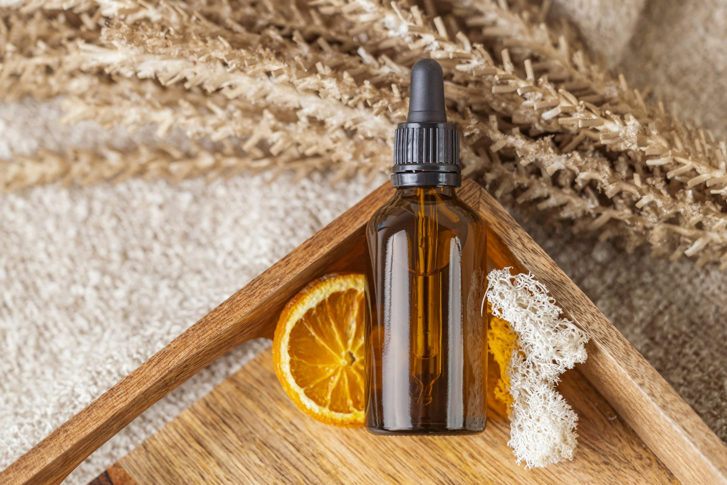 A brown glass dropper bottle placed on a wooden tray, surrounded by dried orange slices and white lichen, with textured beige and patterned background.