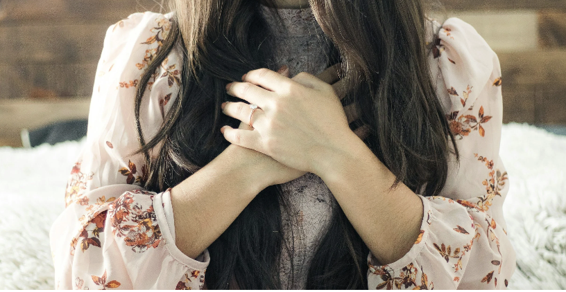 A woman with long dark hair wearing a floral blouse, holding her hands over her chest, sitting on a bed with a white, fluffy blanket.