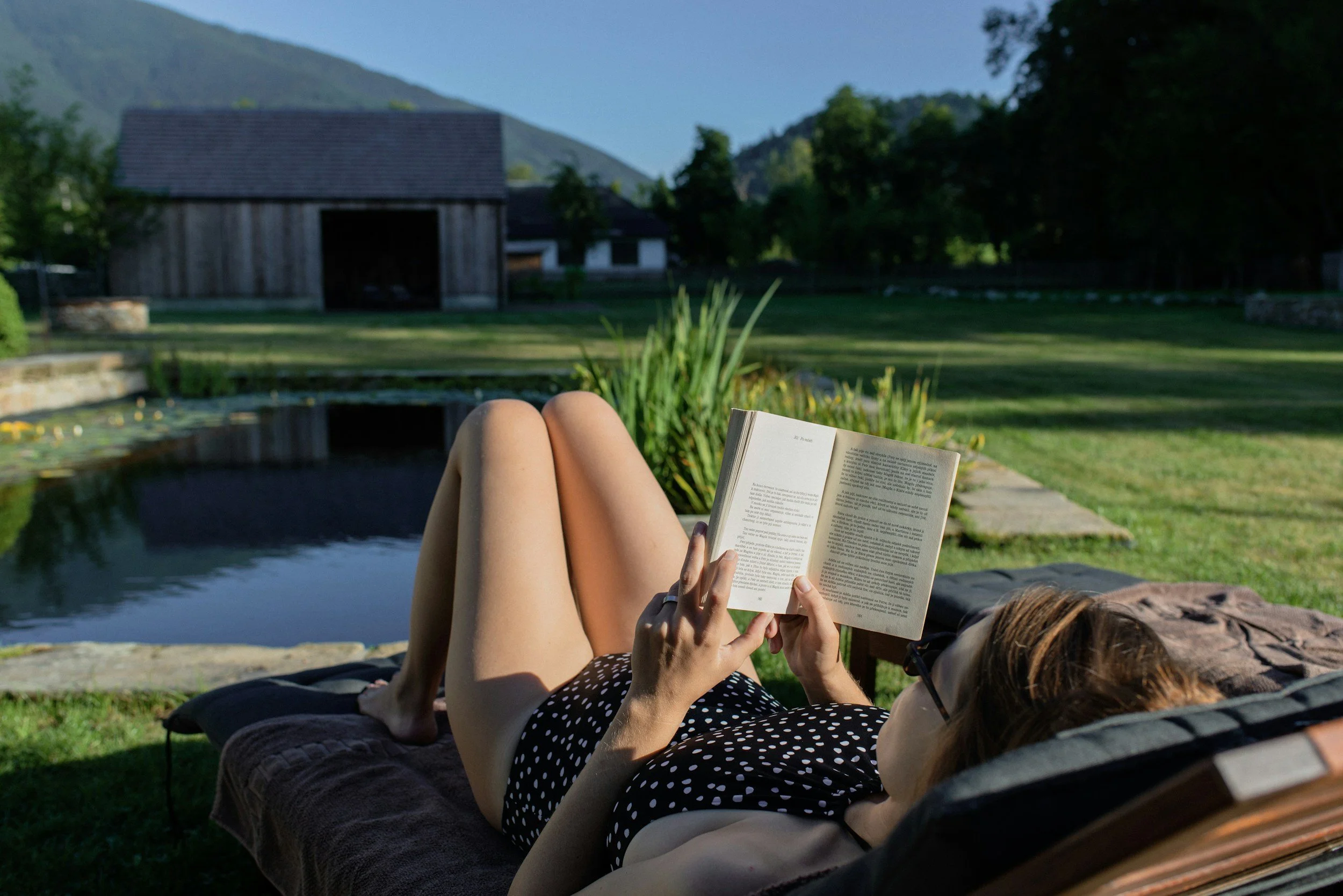 woman sitting on a lounge chair reading a book in front of a pond