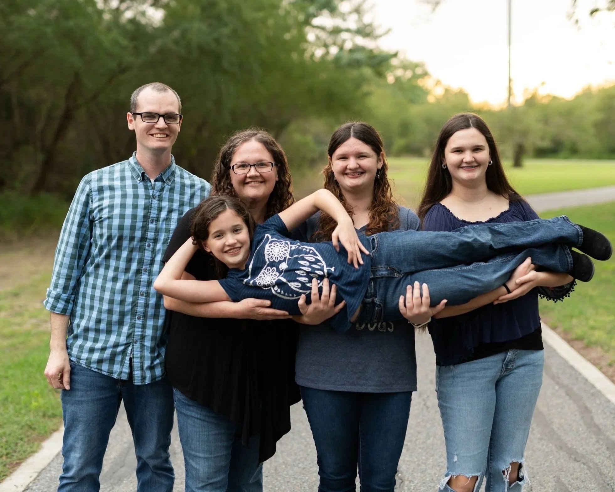 A group of six people, five women and one man, standing outdoors on a path with greenery in the background. One woman is lying across the arms of others, smiling, and wearing a blue top with white floral patterns. The group is smiling and appears happy.