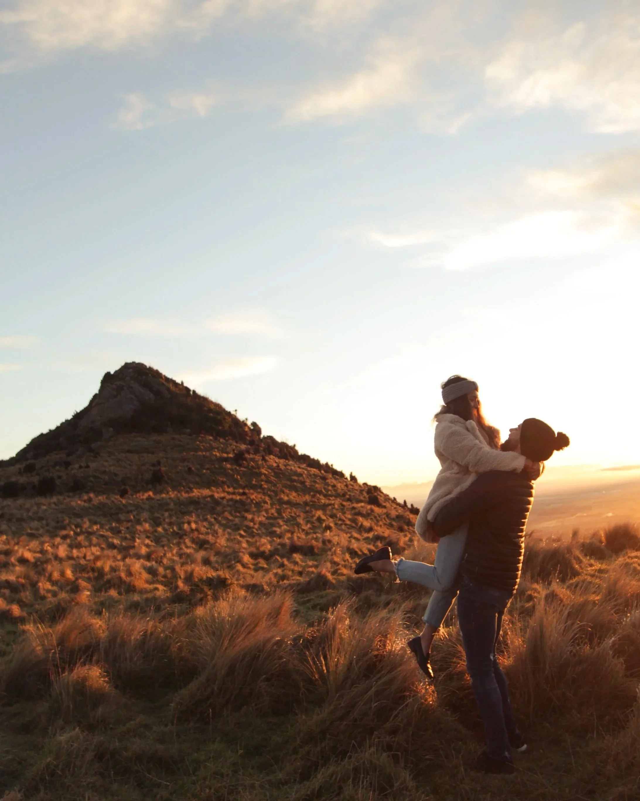 A couple is outdoors during sunset, with the woman giving the man a piggyback ride on a grassy hillside with a mountain in the background.