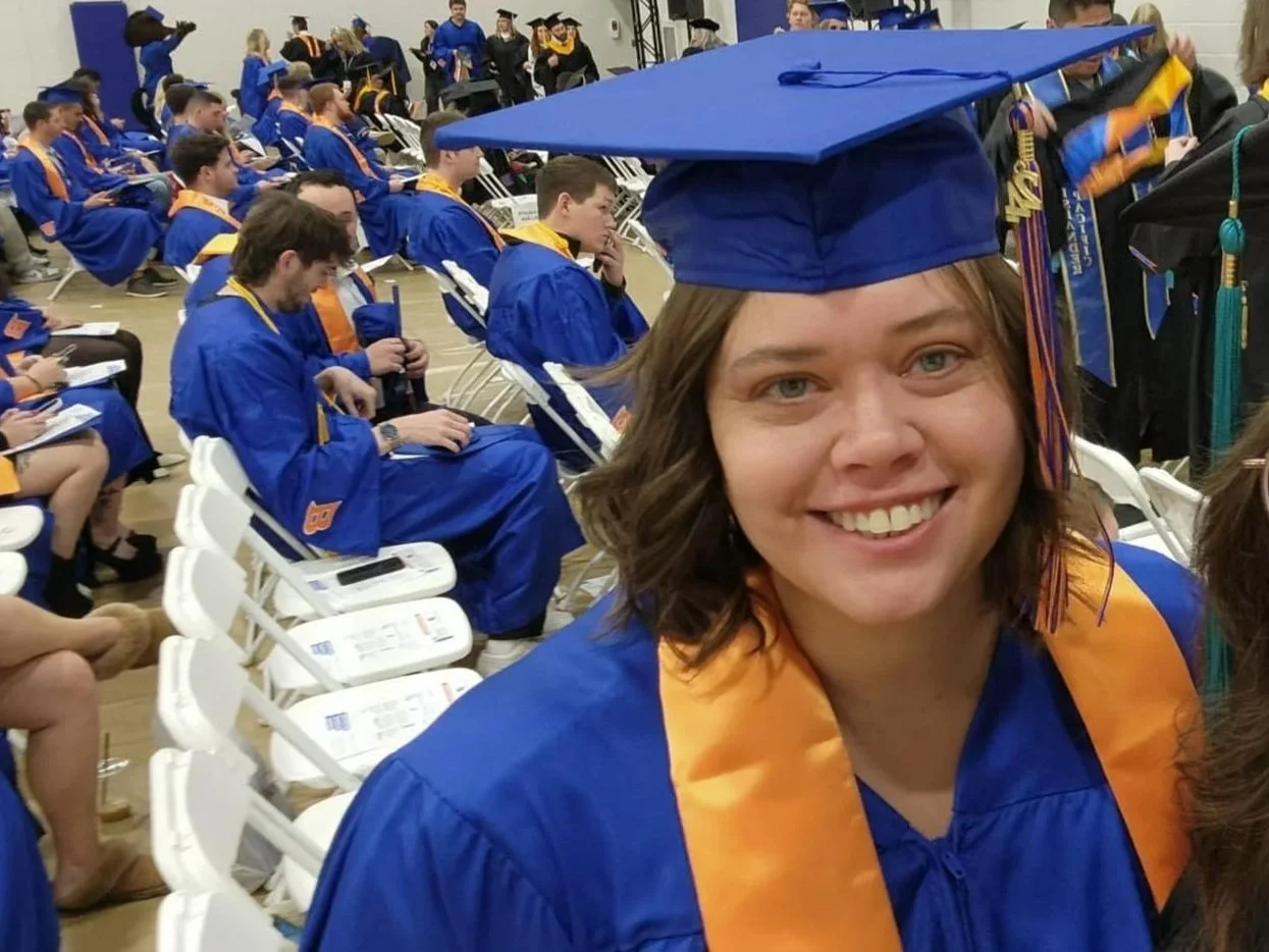 A woman in a blue graduation cap and gown with an orange stole smiling at the camera, at a graduation ceremony with other graduates seated behind her.