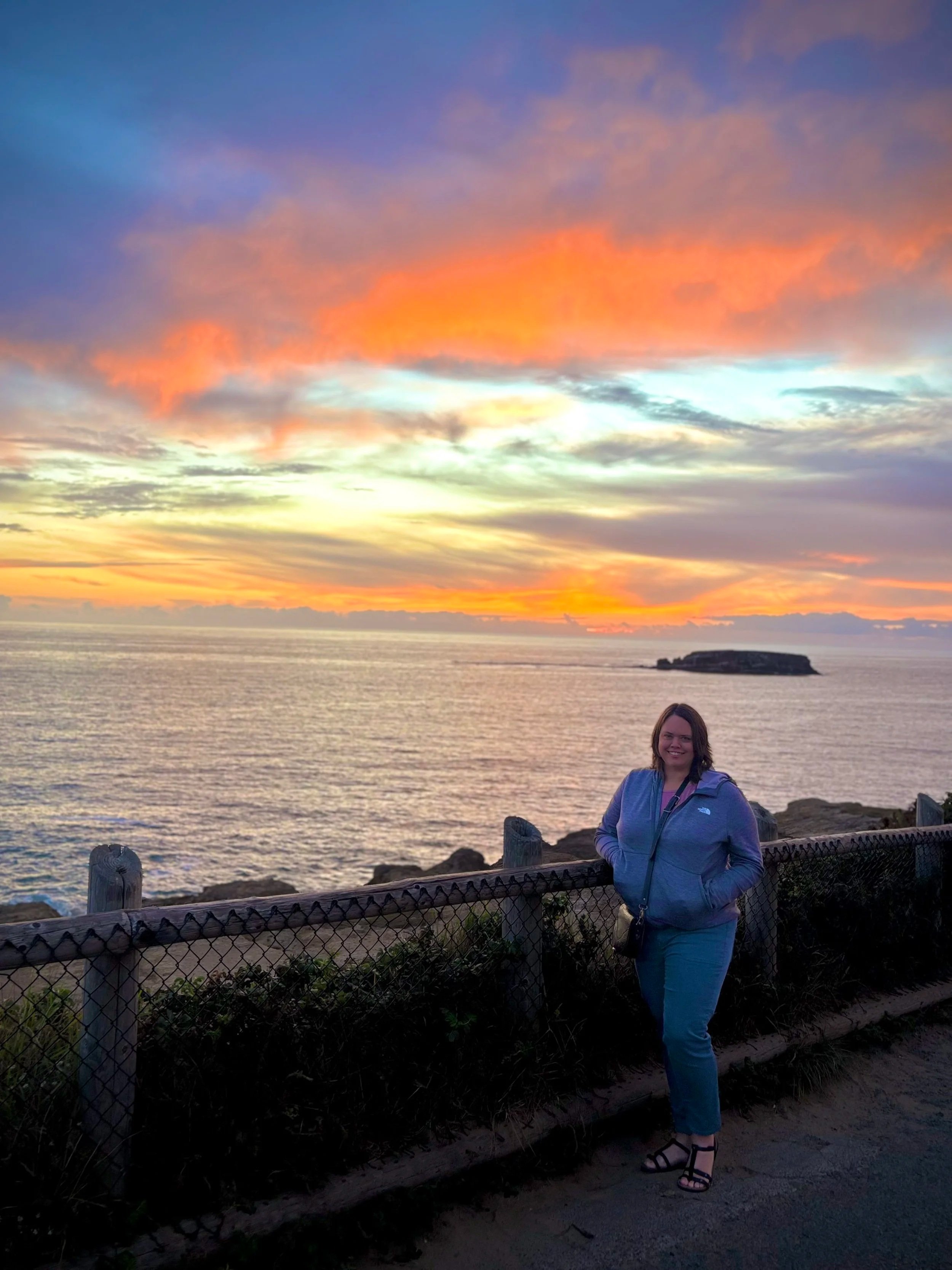 A woman standing outdoors by a fence, smiling, with a sunset over the ocean in the background, displaying colorful clouds in the sky.