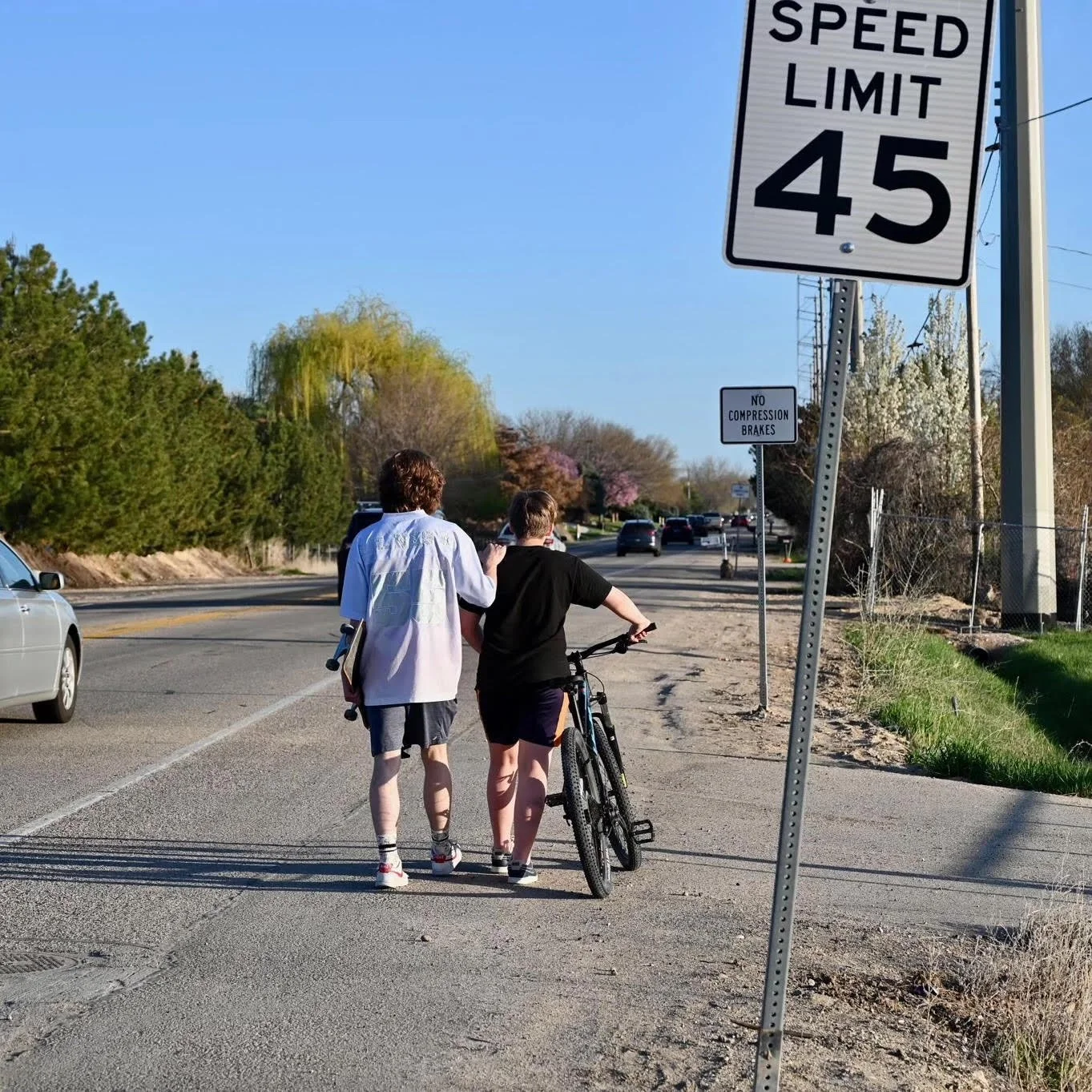 Two people, one with a bicycle, walking on a sidewalk beside a road with cars. There are speed limit signs and a sign that says 'No Compression Brakes'.