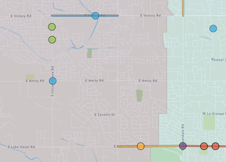 A digital map showing a transportation route with colored circles representing stops along the path. The route includes blue, green, orange, purple, and red stops across a suburban area with streets and some parks or green spaces.