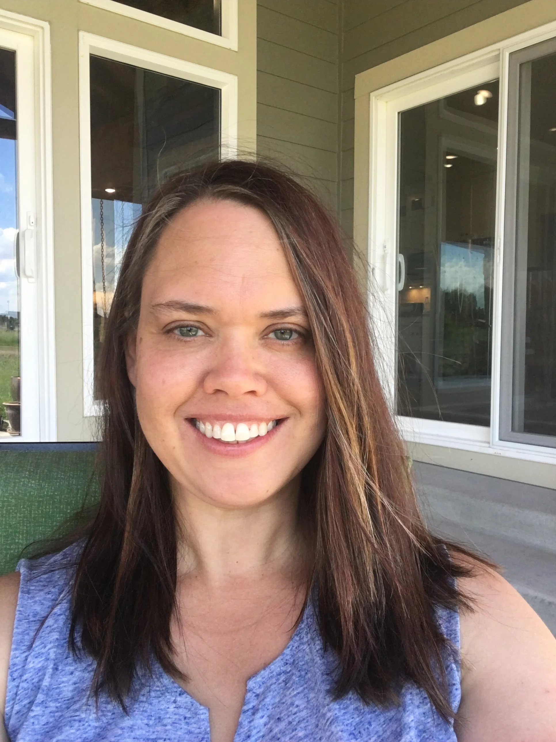 A woman with brown hair, blue eyes, and a bright smile sitting outside in front of a house with large windows and sliding glass doors.
