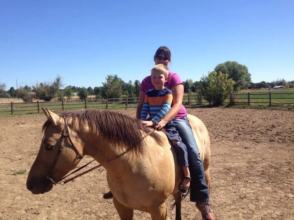 Marisa and a young boy riding a light tan horse in an outdoor arena with a wooden fence, trees, and clear blue sky in the background.