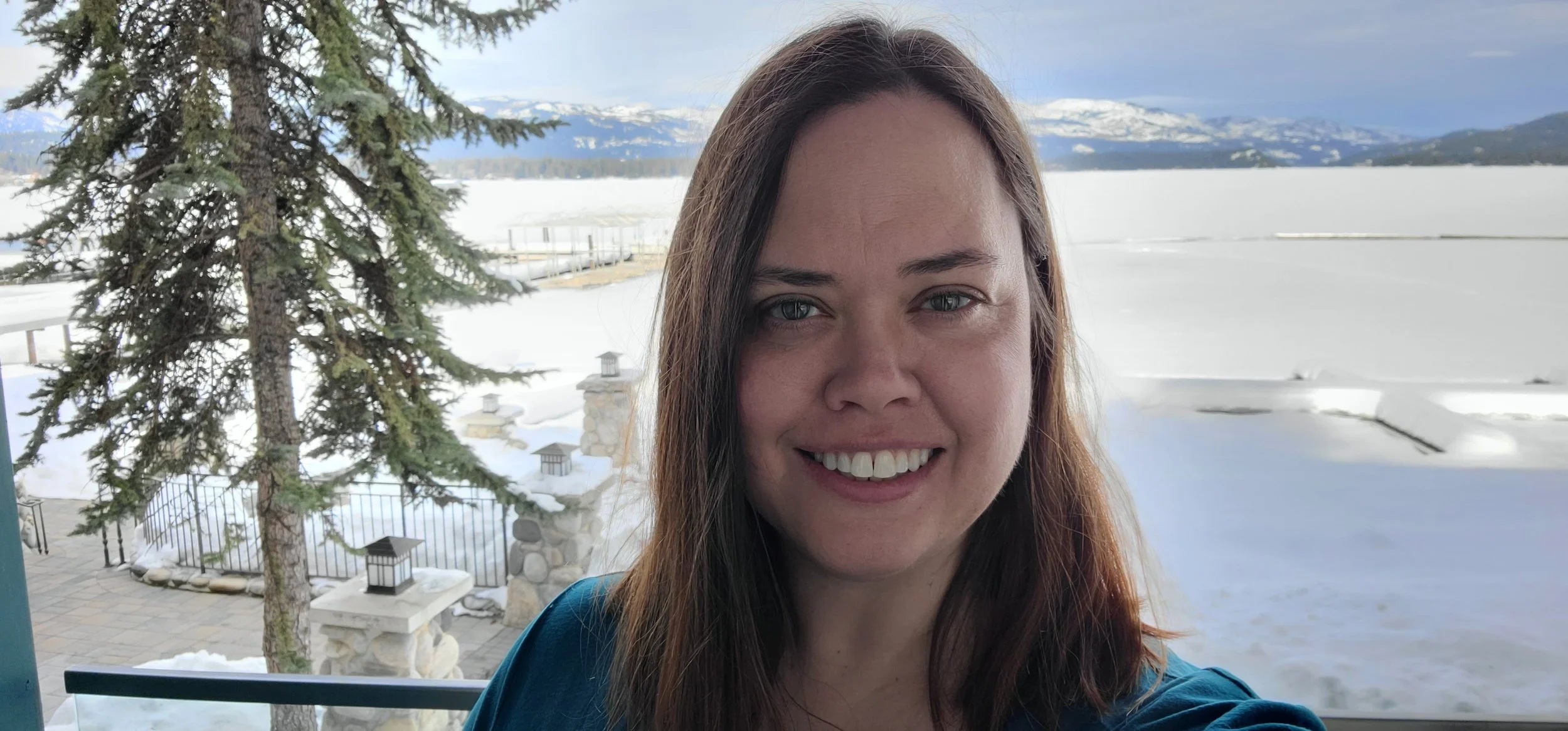 Marisa  with brown hair and blue eyes taking a selfie outdoors with a snowy landscape and mountains in the background.