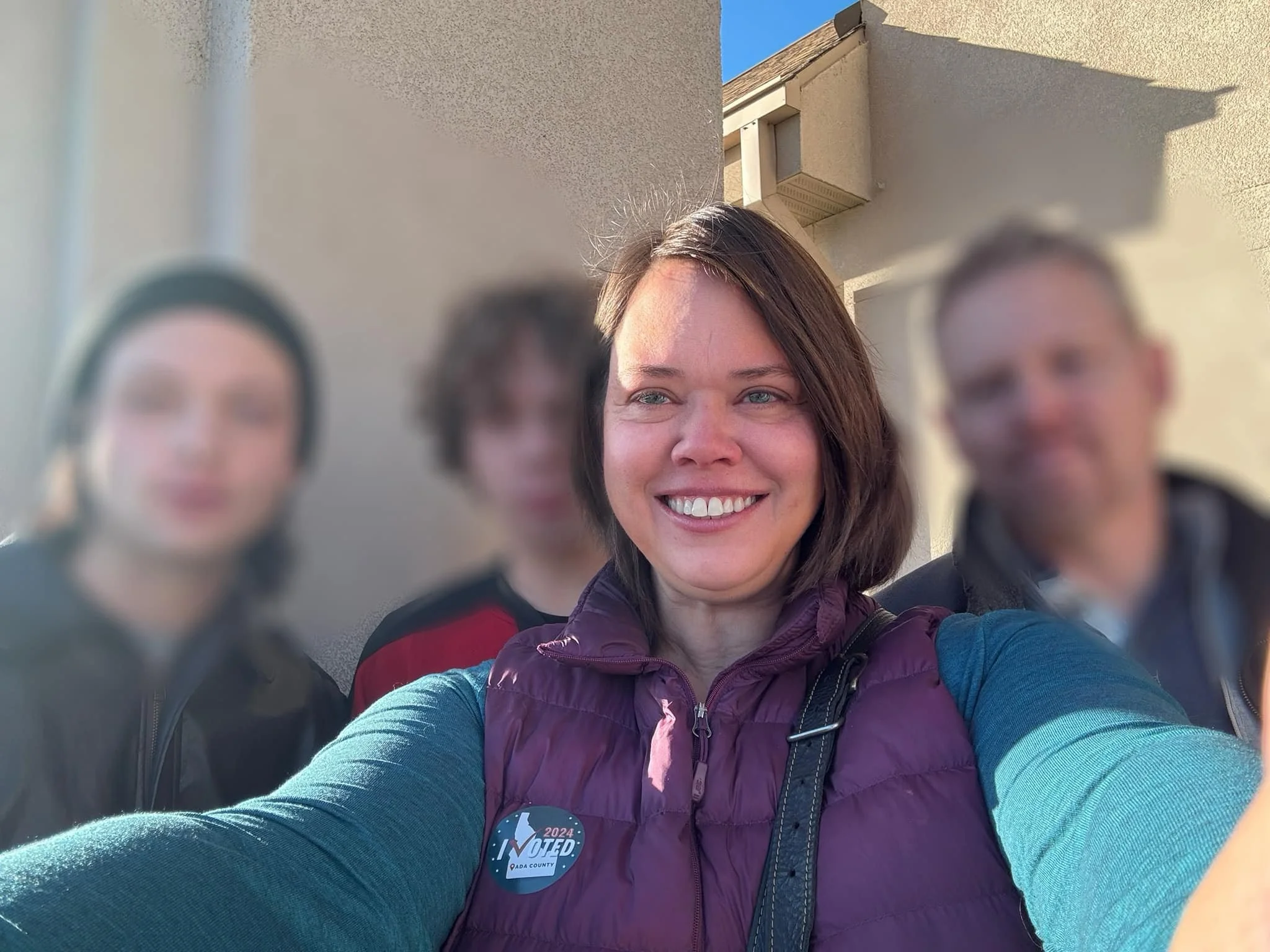 A woman taking a selfie with four people in the background, with the woman smiling at the camera. The others are blurred, and one person is wearing a badge that says "2024 VOTED DADE COUNTY." The background shows a beige building and a clear blue sky