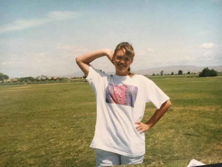 A young girl standing in a grassy field, smiling with one hand on her hip and the other behind her head, under a partly cloudy sky.