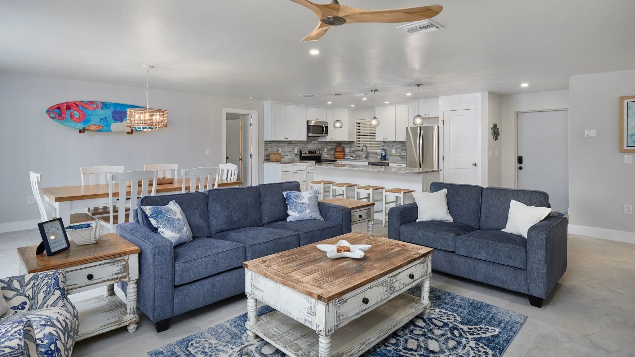 Living room with two blue sofas, white distressed coffee table, and navy blue and white area rug. Open kitchen with white cabinets, gray backsplash, stainless steel appliances, and white island with bar stools. Wall art of a colorful surfboard, dining table with white chairs, ceiling fan, and a bright, modern decor style.
