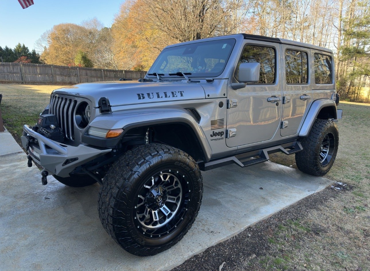 A silver Jeep Wrangler parked on a concrete pad in a backyard during autumn, with a wooden fence and trees with fall foliage in the background.
