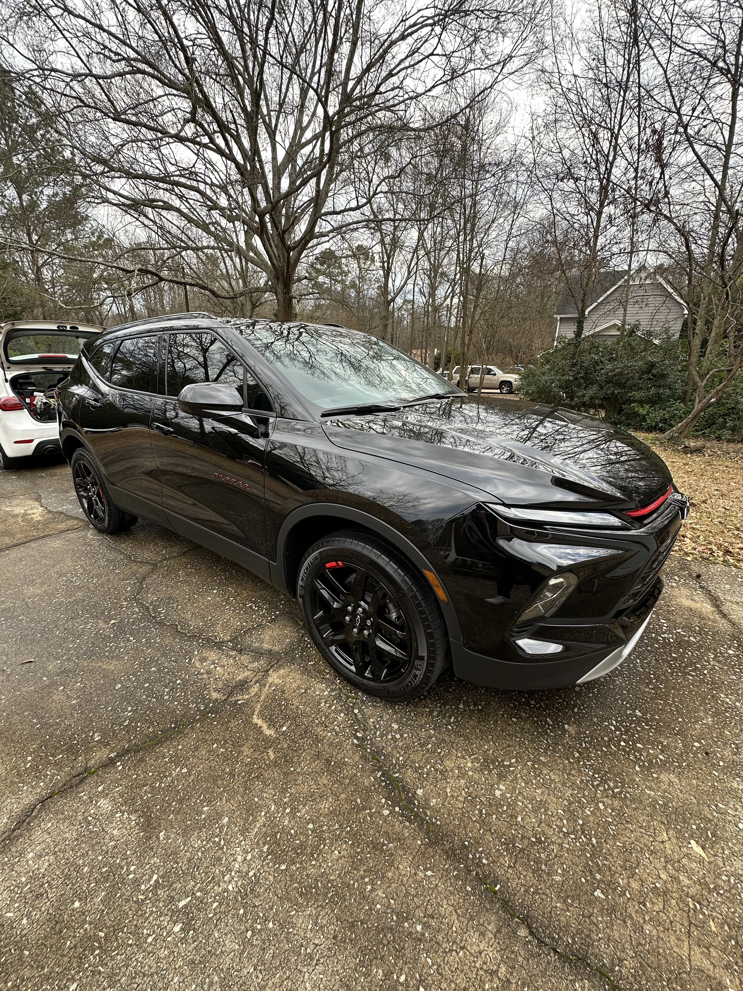 A black SUV parked on a cracked concrete driveway in front of a leafless tree and suburban houses, with another white car in the background.