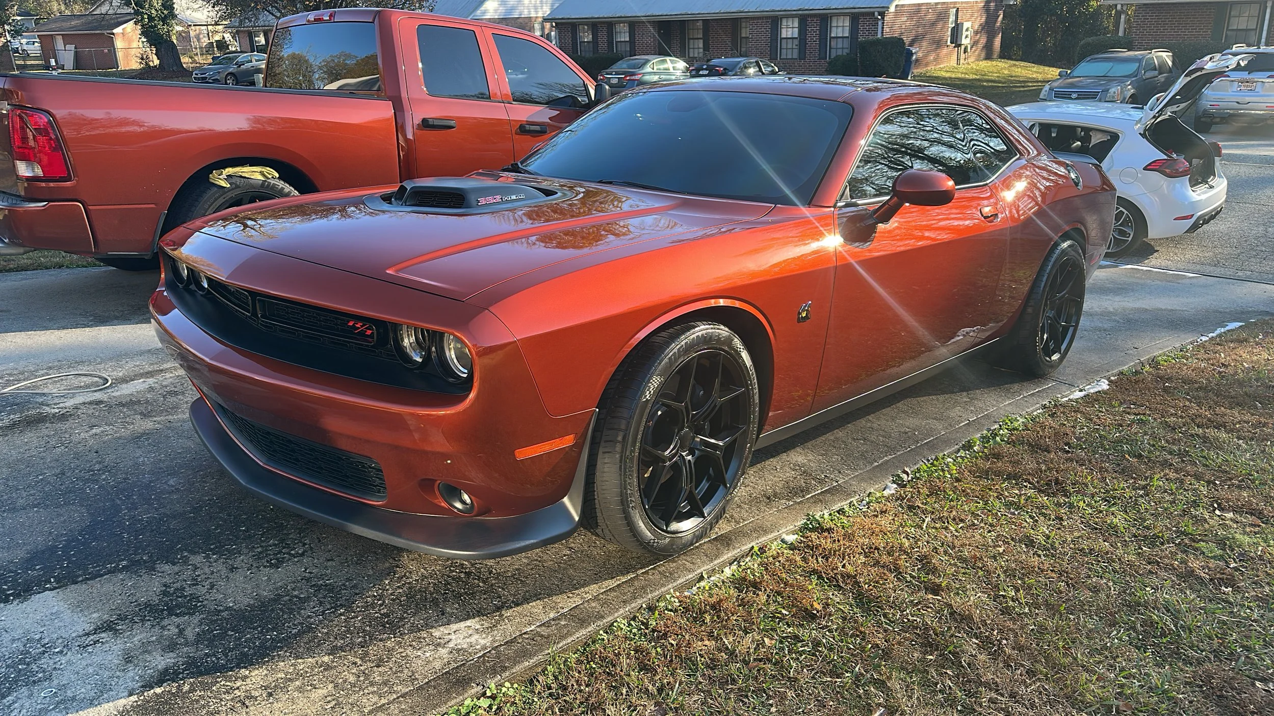 A red Dodge Challenger muscle car parked in a parking lot next to a red pickup truck and a white car with its trunk open.
