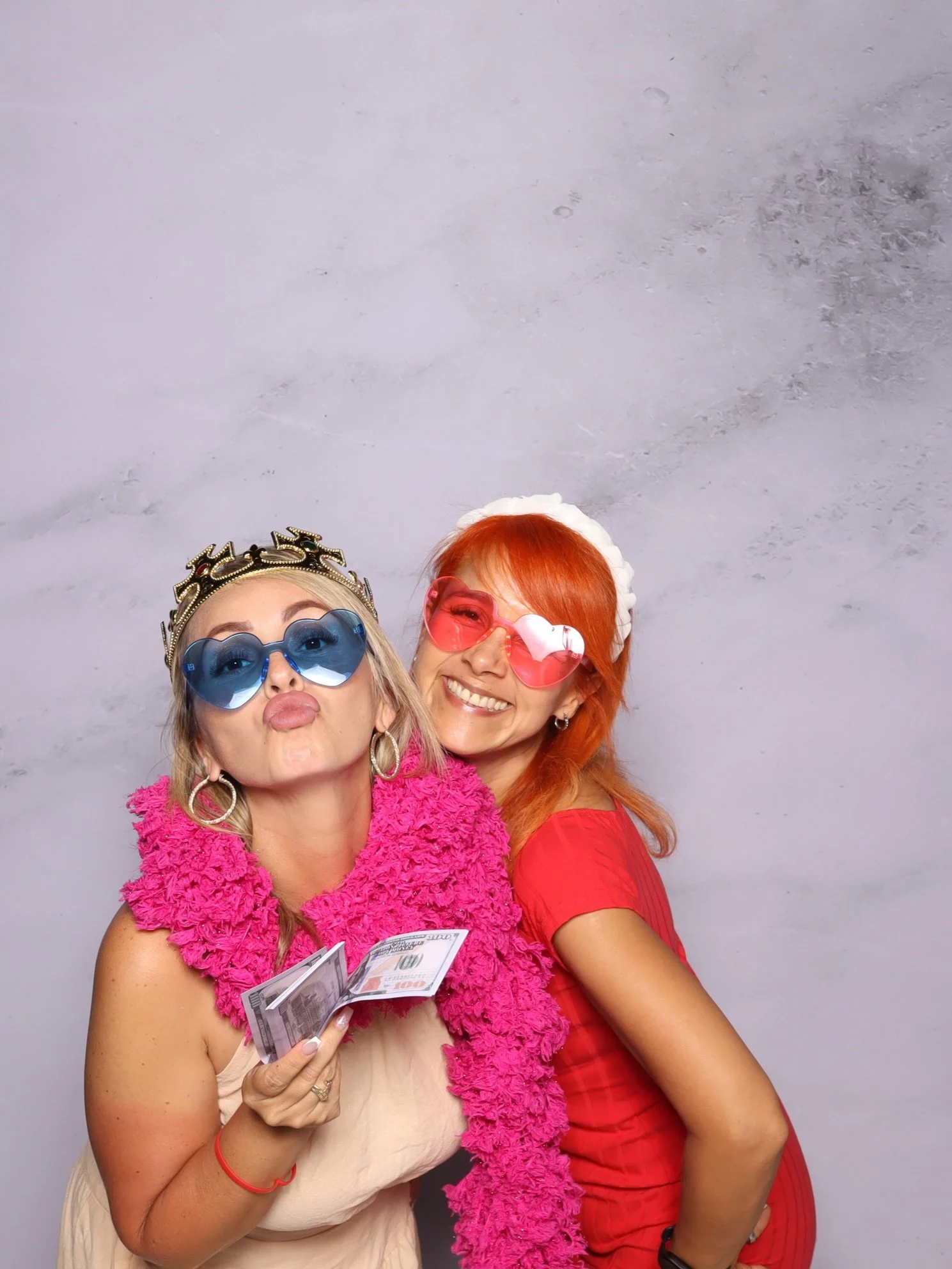 Two women in colorful outfits and sunglasses posing for a fun photo, one holding cash, against a plain background.