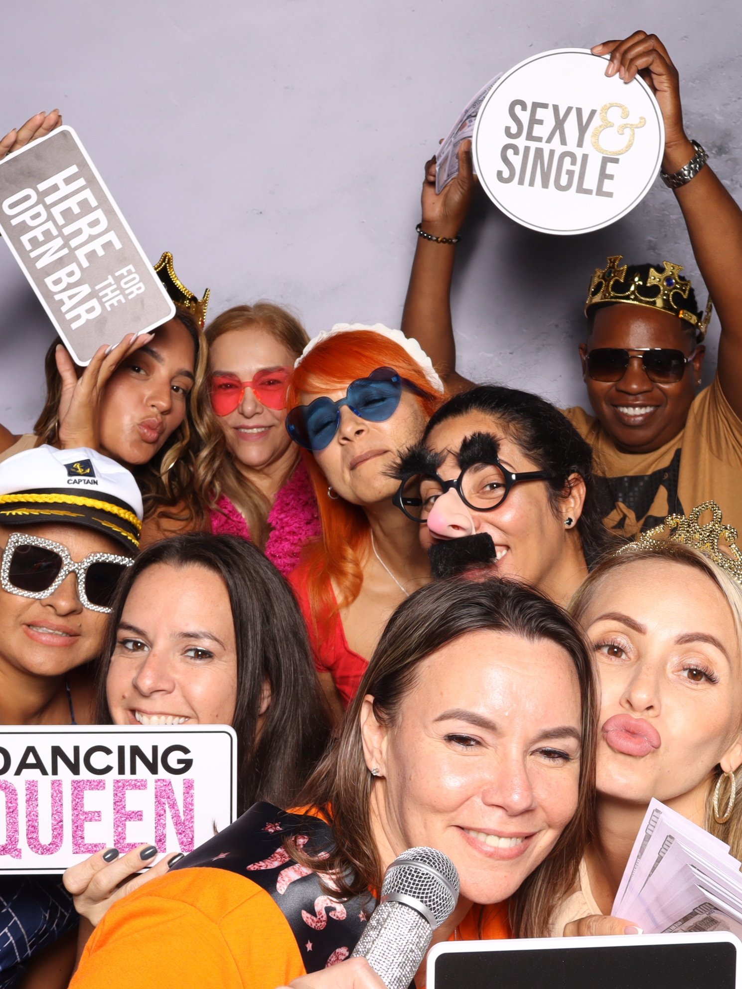 Group of people at a party with props like crowns, glasses, and signs that read 'Sexy & Single,' 'Here for the Open Bar,' and 'Dancing Queen.'