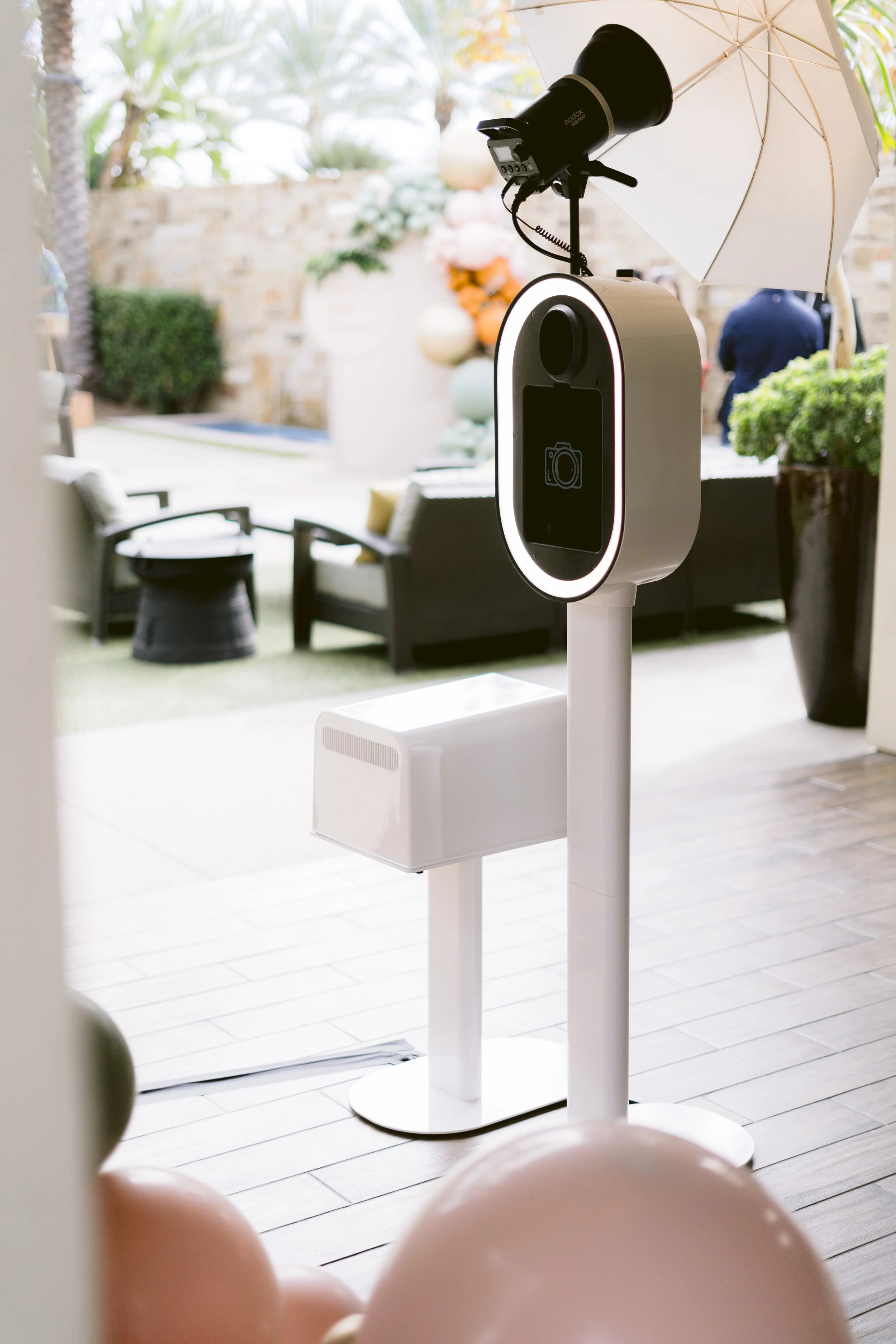 A photo of a photo booth with a camera and light ring, set up outdoors on a patio with balloons, chairs, and a table in the background.