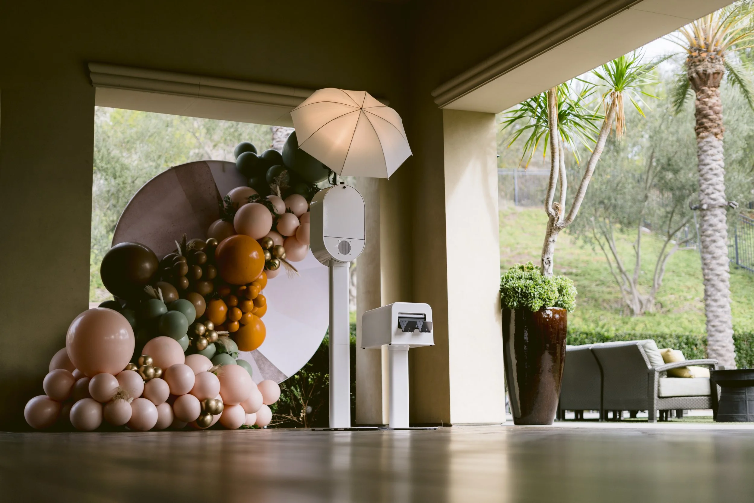 Decorative balloon arrangement with an umbrella and photo booth props on a porch, with outdoor trees and seating visible in the background.