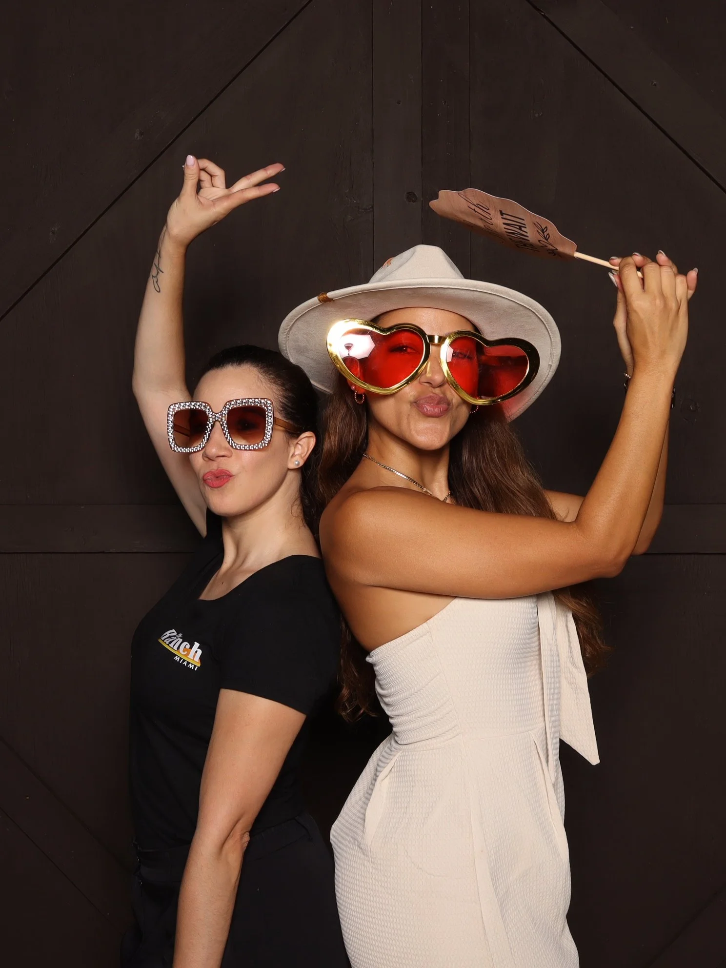 Two women posing playfully against a dark wooden wall, wearing oversized sunglasses and hats, with one holding a sign and the other making a peace sign.