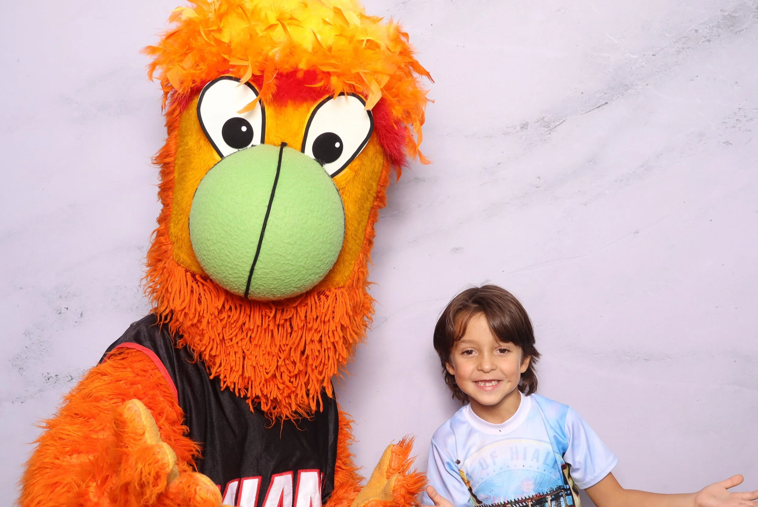 A young boy smiling and pointing with his right hand next to a large mascot costume of a lion with an orange mane, green nose, and black jersey, standing against a light-colored wall.