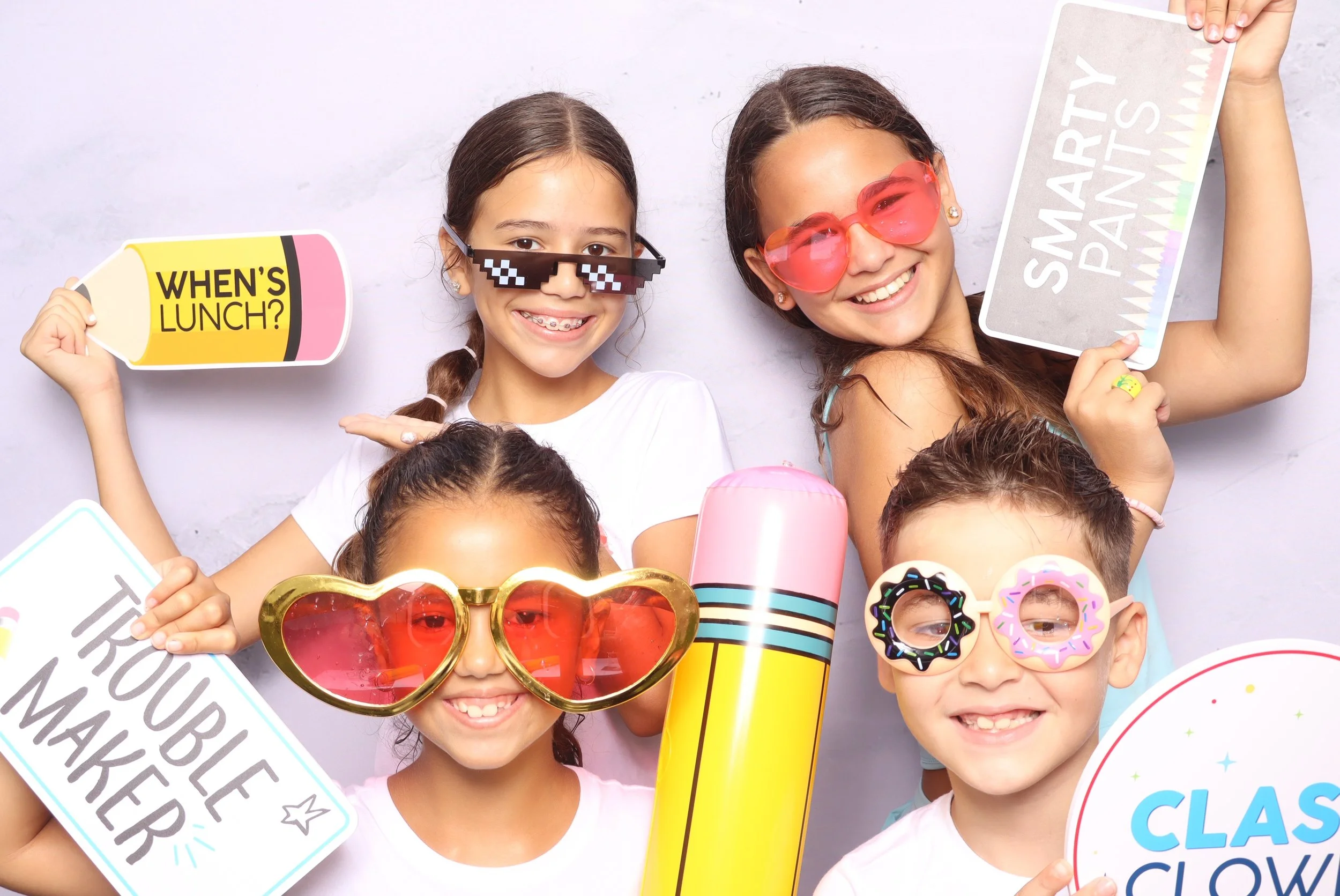 Group of five kids celebrating with playful props, including oversized glasses, signs, a giant pencil, and a large colorful lollipop.
