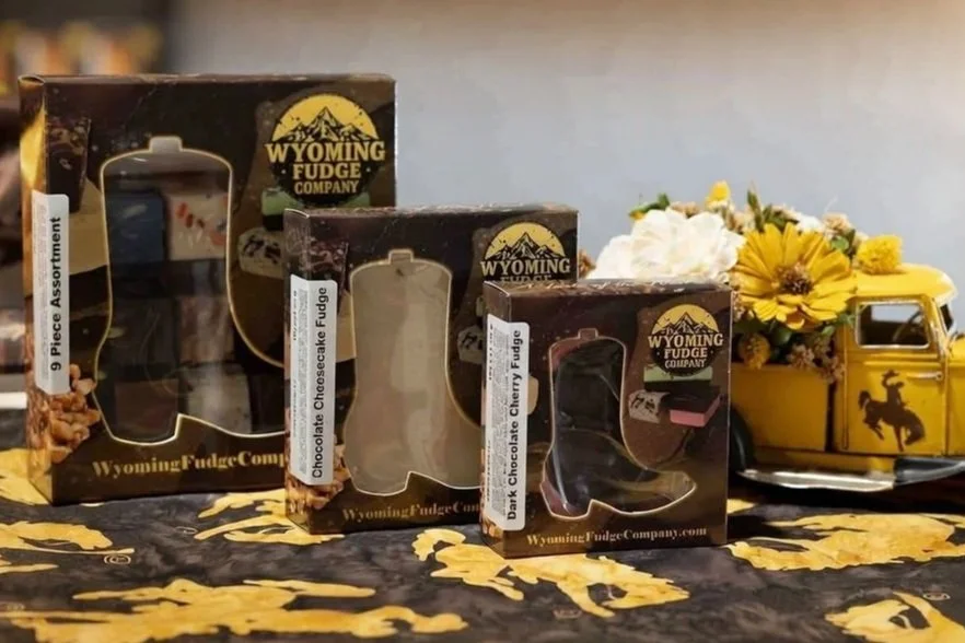 Three boxes of Wyoming Fudge Company fudge flavors (Chocolate Cheesecake, Dark Chocolate Cherry, and another) with a yellow decorative truck filled with white and yellow flowers in the background.