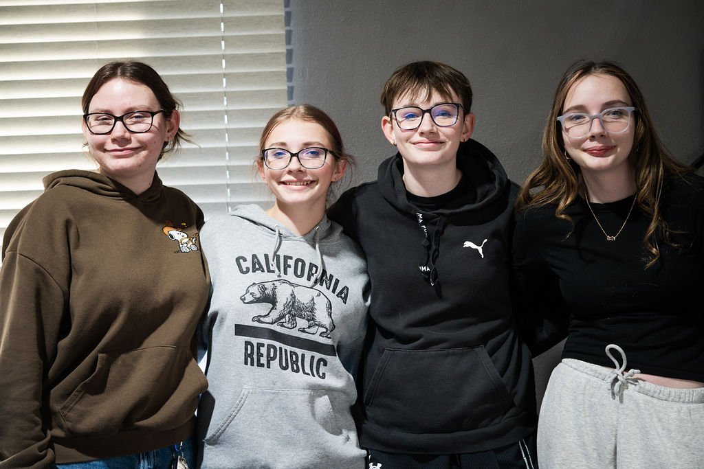 Four teenagers standing together indoors in front of a window with blinds, smiling at the camera.