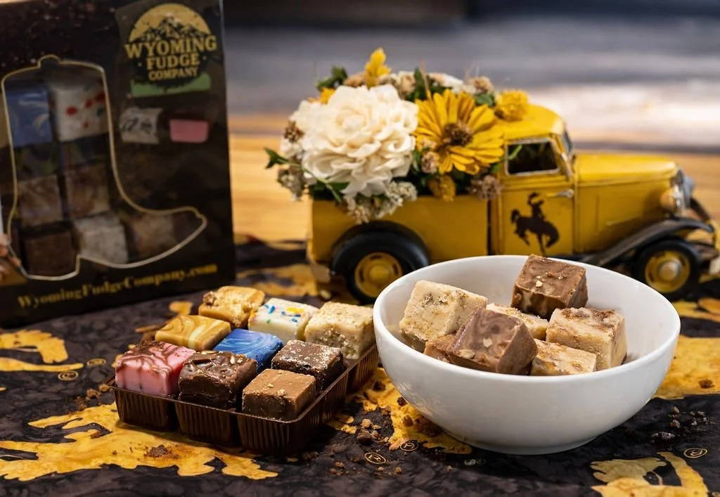 Assorted flavored fudge pieces in a black tray and white bowl placed on a table with a yellow and black tablecloth, a decorative yellow vintage truck filled with white and yellow flowers, and a box of Wyoming Fudge Company chocolates in the backgroun