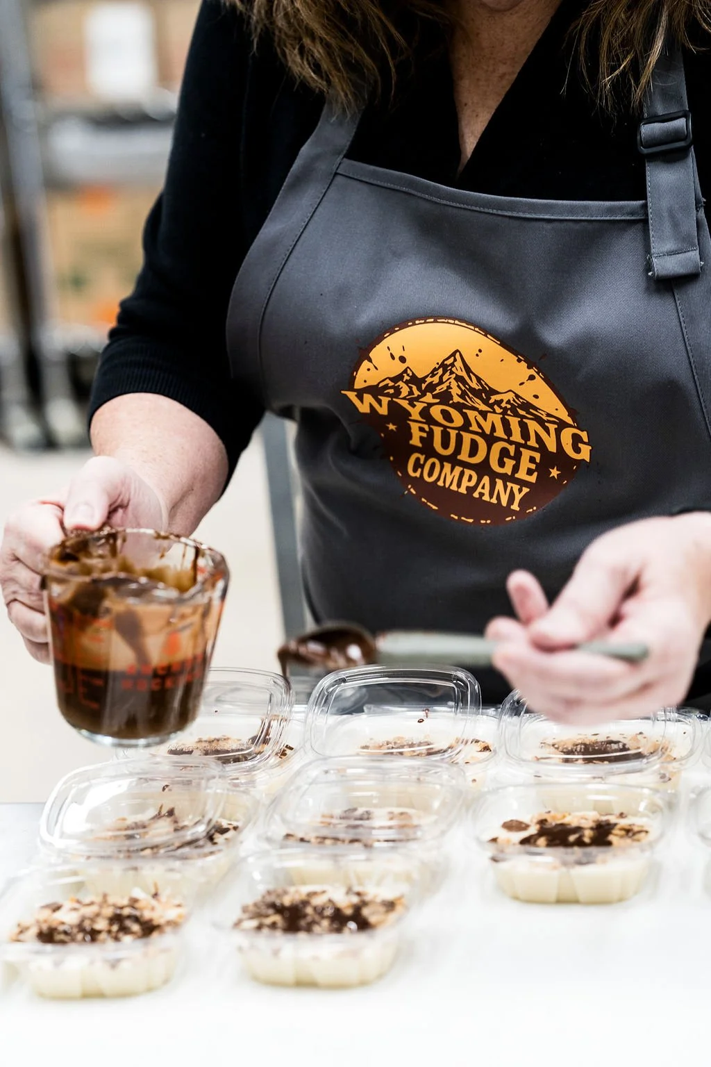 Person wearing a Wyoming Fudge Company apron preparing dessert cups with chocolate topping in a kitchen or shop environment.