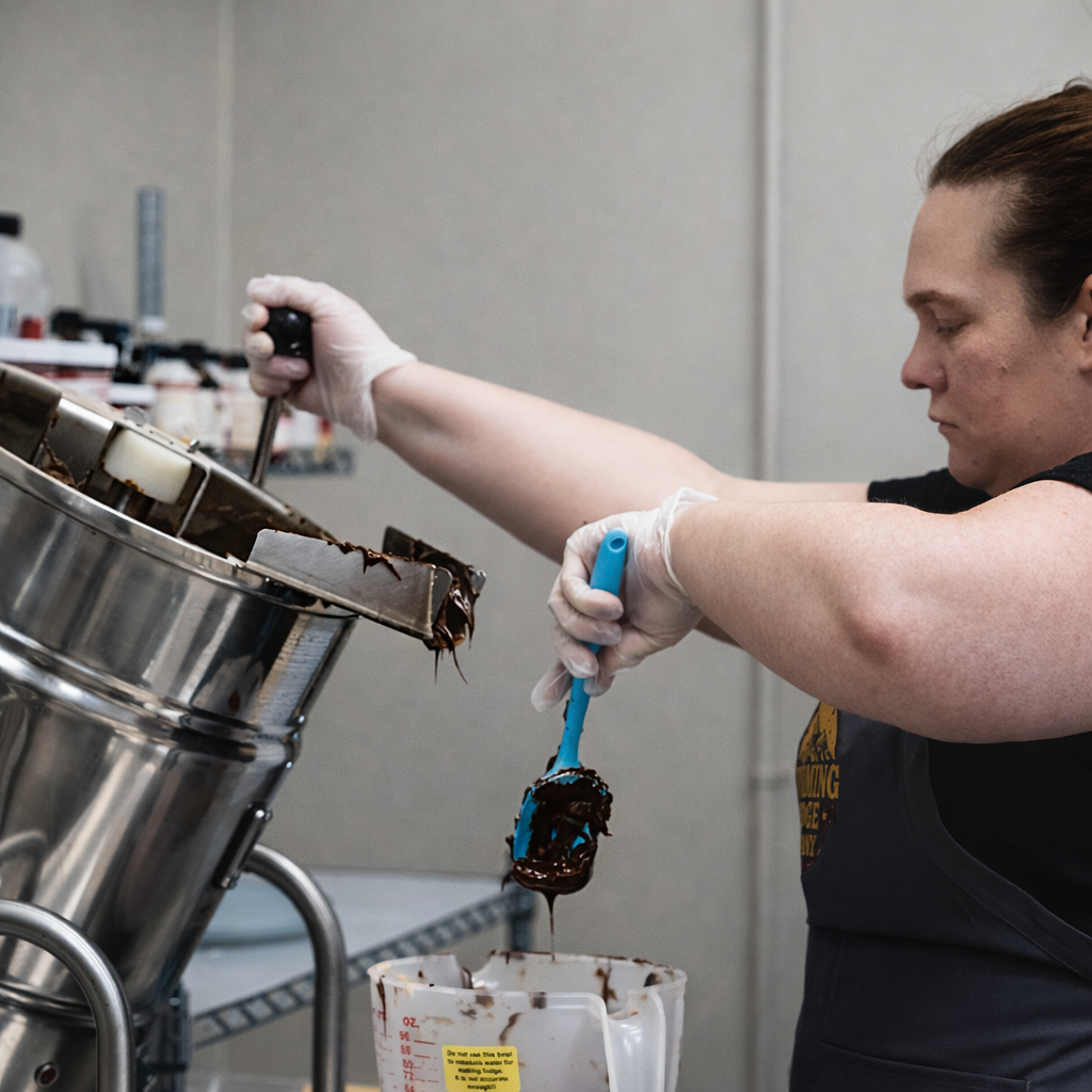 A person using a spatula to scrape melted chocolate from a machine into a container, wearing gloves, in a kitchen or food production setting.
