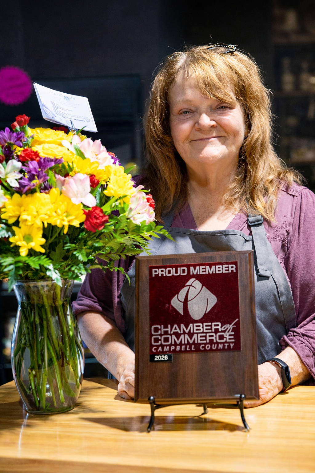 A woman with reddish hair standing behind a wooden table with a bouquet of colorful flowers and a sign reading 'Proud Member Chamber of Commerce Campbell County.'