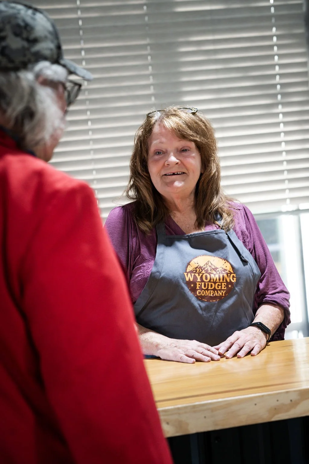 A woman with brown hair and glasses on her head, wearing a Wyoming Fudge Company apron, is smiling and talking to a person in a red jacket in a store or shop with closed window blinds in the background.