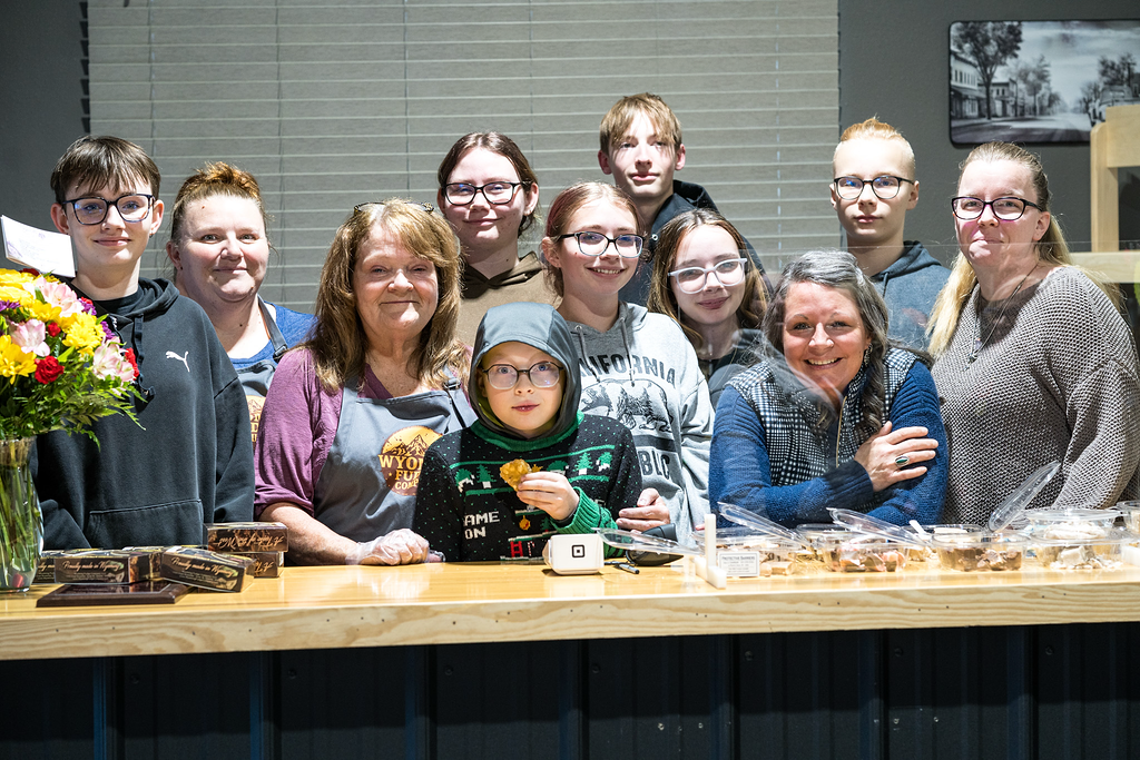 Group of children and adults gathered behind a wooden counter with baked goods, smiling in a bakery or community event setting.