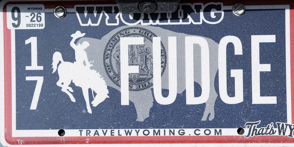Wyoming license plate featuring a cowboy riding a bucking bronco, a silhouette of a wolf, and the words 'Wyoming', 'Fudge', and the website 'travelwyoming.com'.