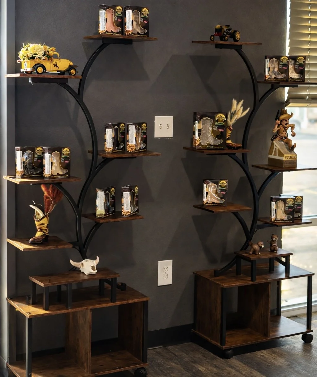 Decorative display shelf with cowboy-themed figurines, toy boots, and vintage-style toy vehicles, with a dark wall and a window with blinds in the background.