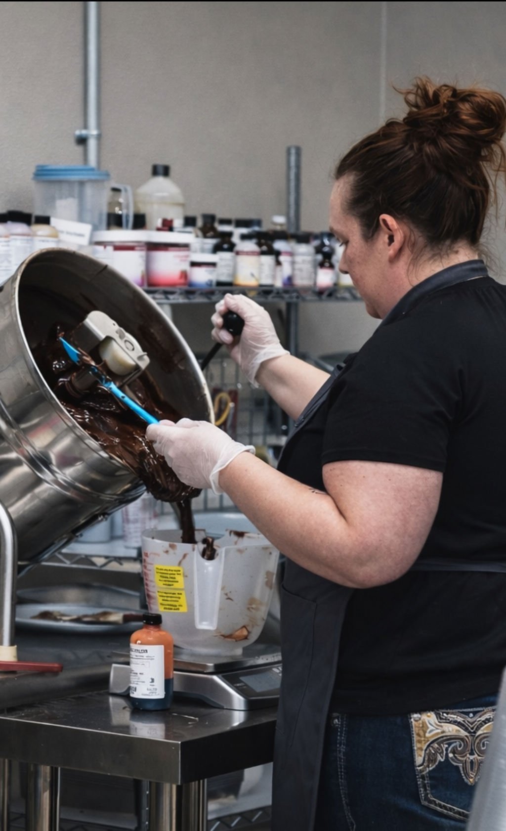 A person pouring melted chocolate from a large metal container into a white container. The person is wearing gloves and a black shirt, working in a kitchen or laboratory setting with shelves of bottles and jars behind them.