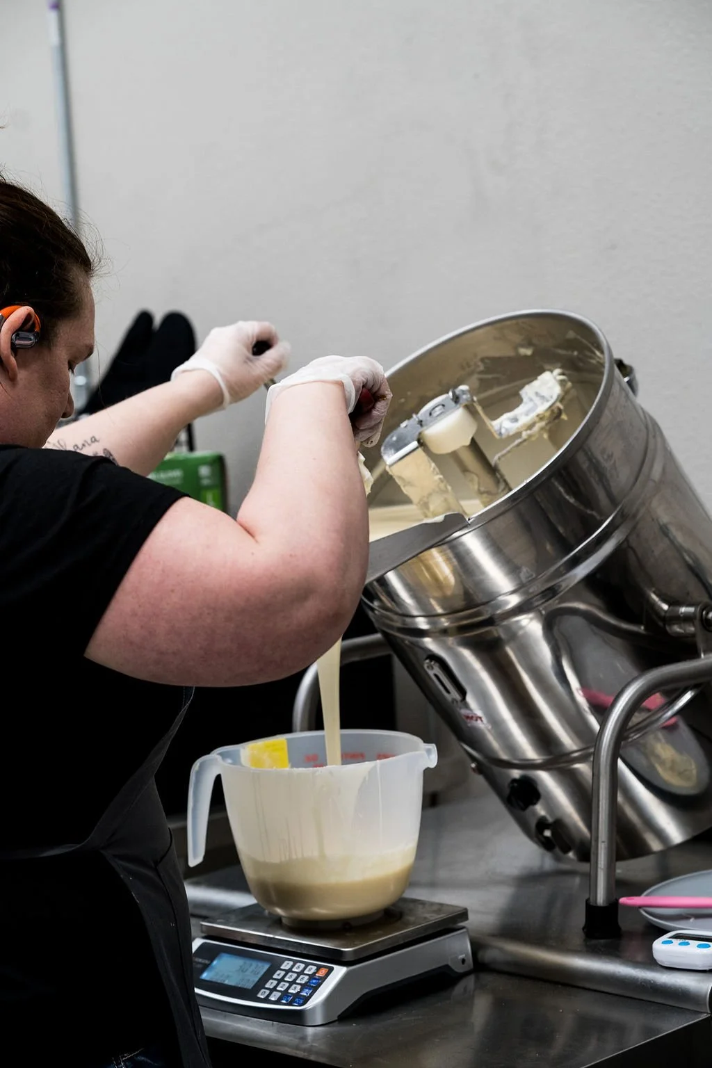 A person pours a cream mixture from a metal industrial mixer into a measuring cup on a scale in a commercial kitchen.