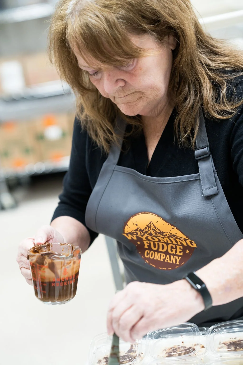 A woman wearing a grey apron with the logo 'Wyoming Fudge Company' is pouring hot fudge from a spoon into a glass cup.